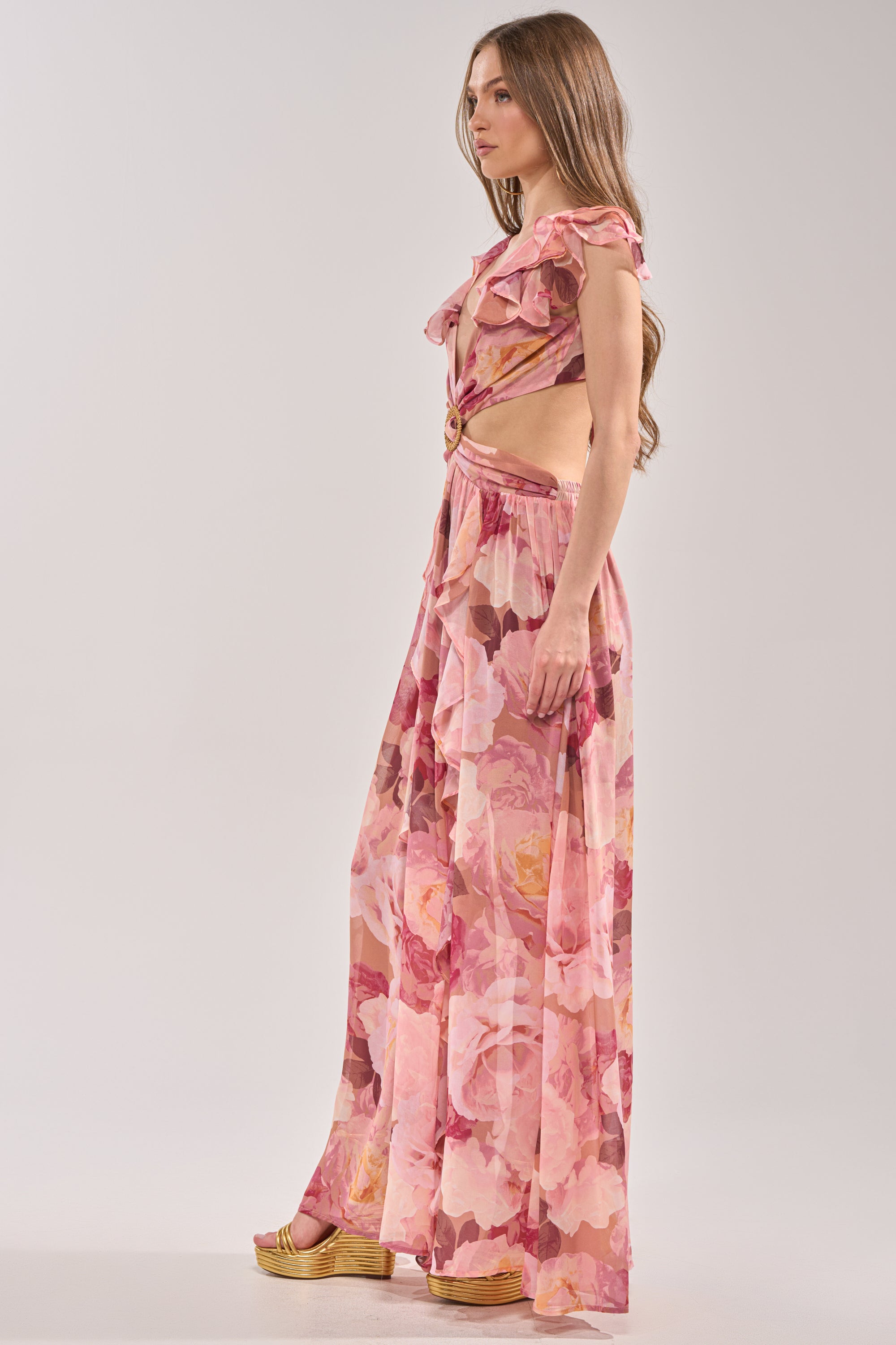 A woman in profile models the LOVE YOU LATER FLORAL MAXI DRESS, a pink chiffon maxi with ruffles and cutout sides. Her long brown hair is loose, and she pairs the flowing floral dress with platform sandals—ideal for spring occasions. 3