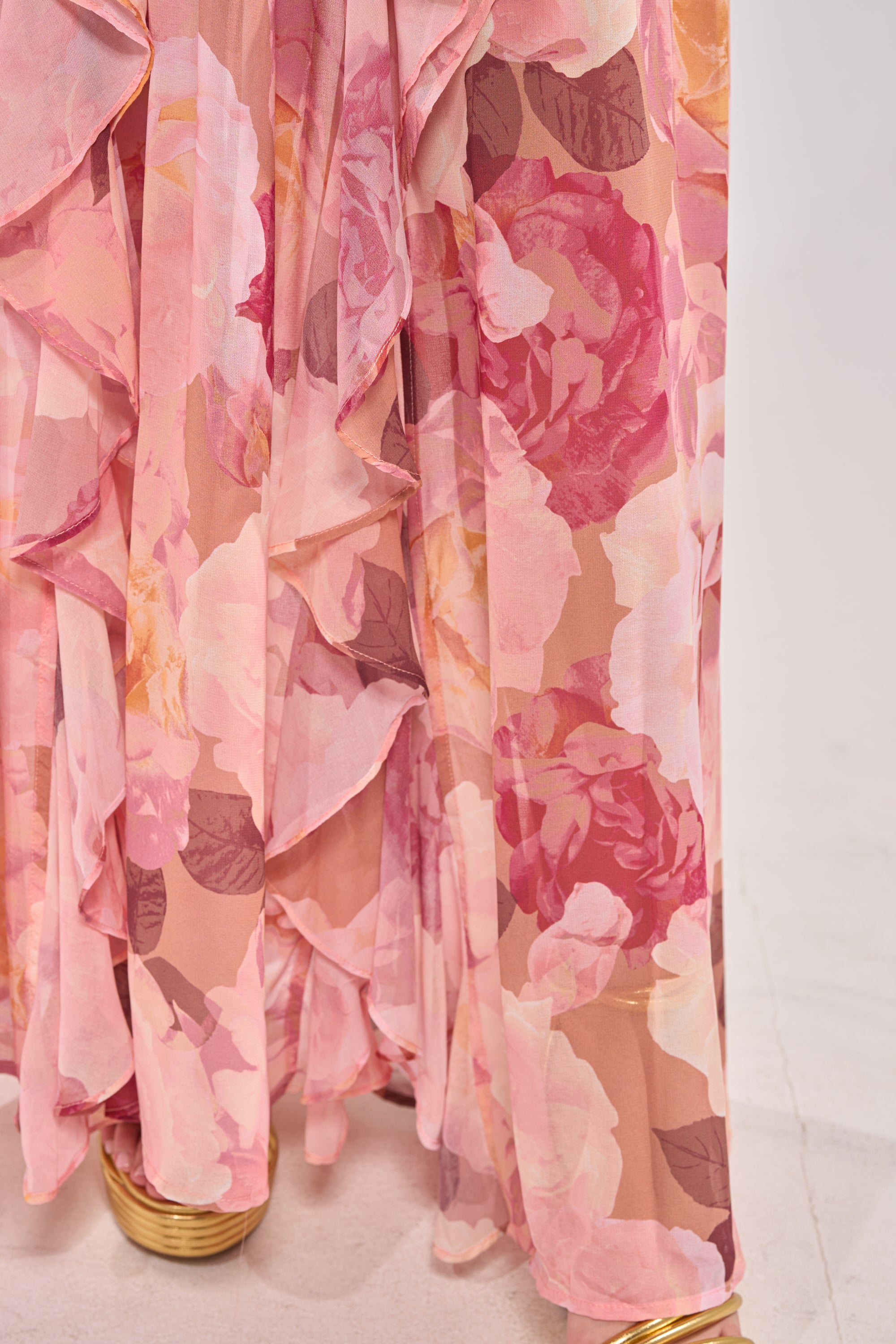A close-up of the LOVE YOU LATER FLORAL MAXI DRESS, a flowing chiffon maxi with large pink and mauve floral patterns—perfect for spring occasions—paired with gold sandals on a light-toned floor. 7