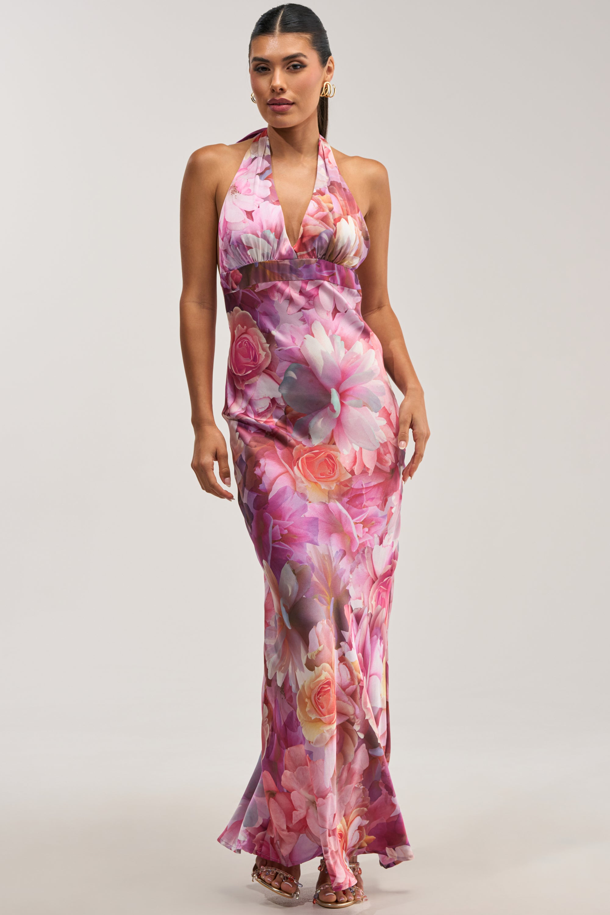 A woman stands against a plain light background, wearing the HOPELESS ROMANTIC FLORAL SATIN MAXI DRESS—a floor-length halter maxi dress adorned with a pink floral pattern. She has long dark hair, gold hoop earrings, and open-toe shoes, making this an elegant choice for a wedding guest outfit.