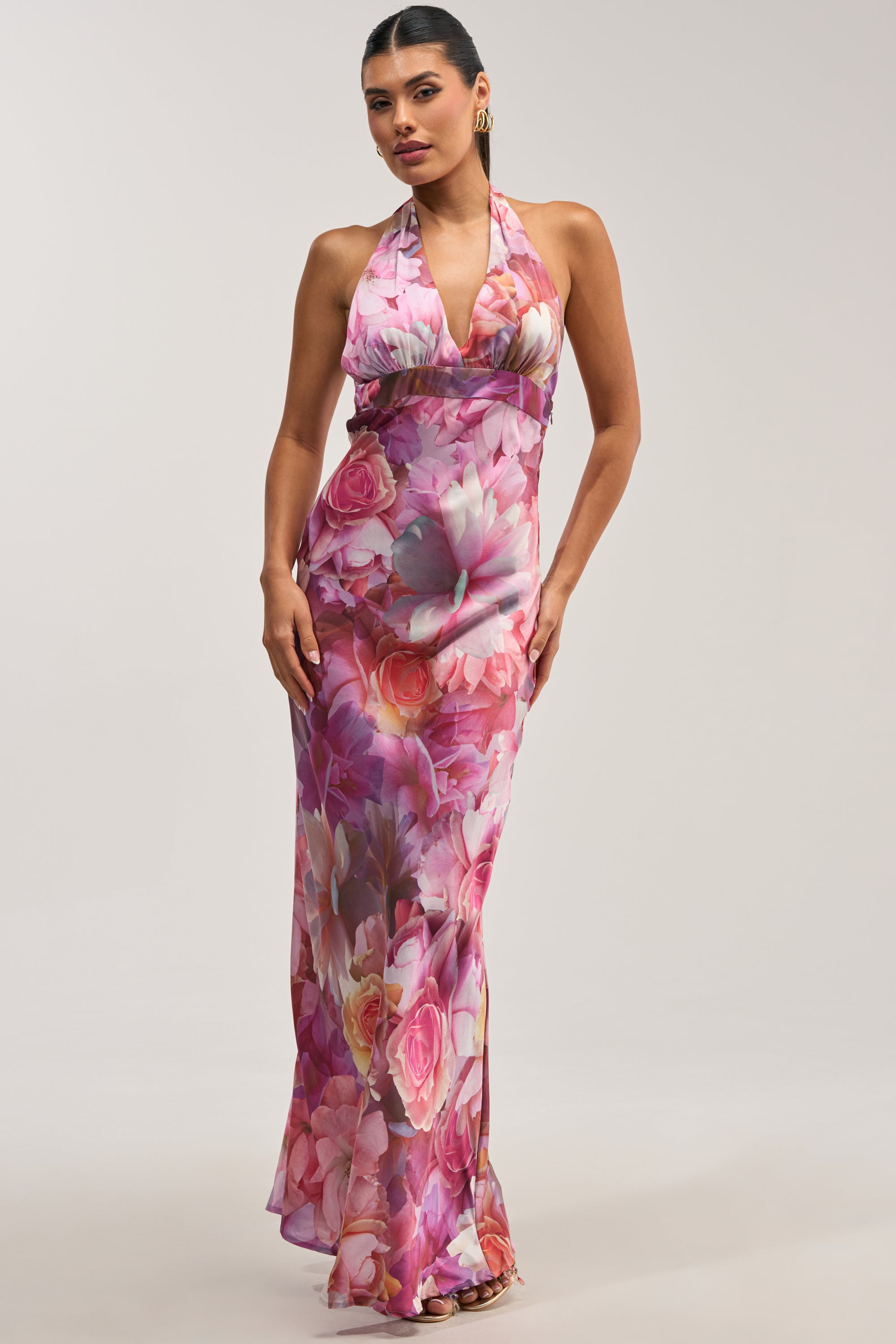 A woman wearing the HOPELESS ROMANTIC FLORAL SATIN MAXI DRESS, a long satin maxi dress featuring a sleeveless halter neckline and a vibrant pink and purple floral print, stands against a plain light background—an elegant choice for wedding guests.