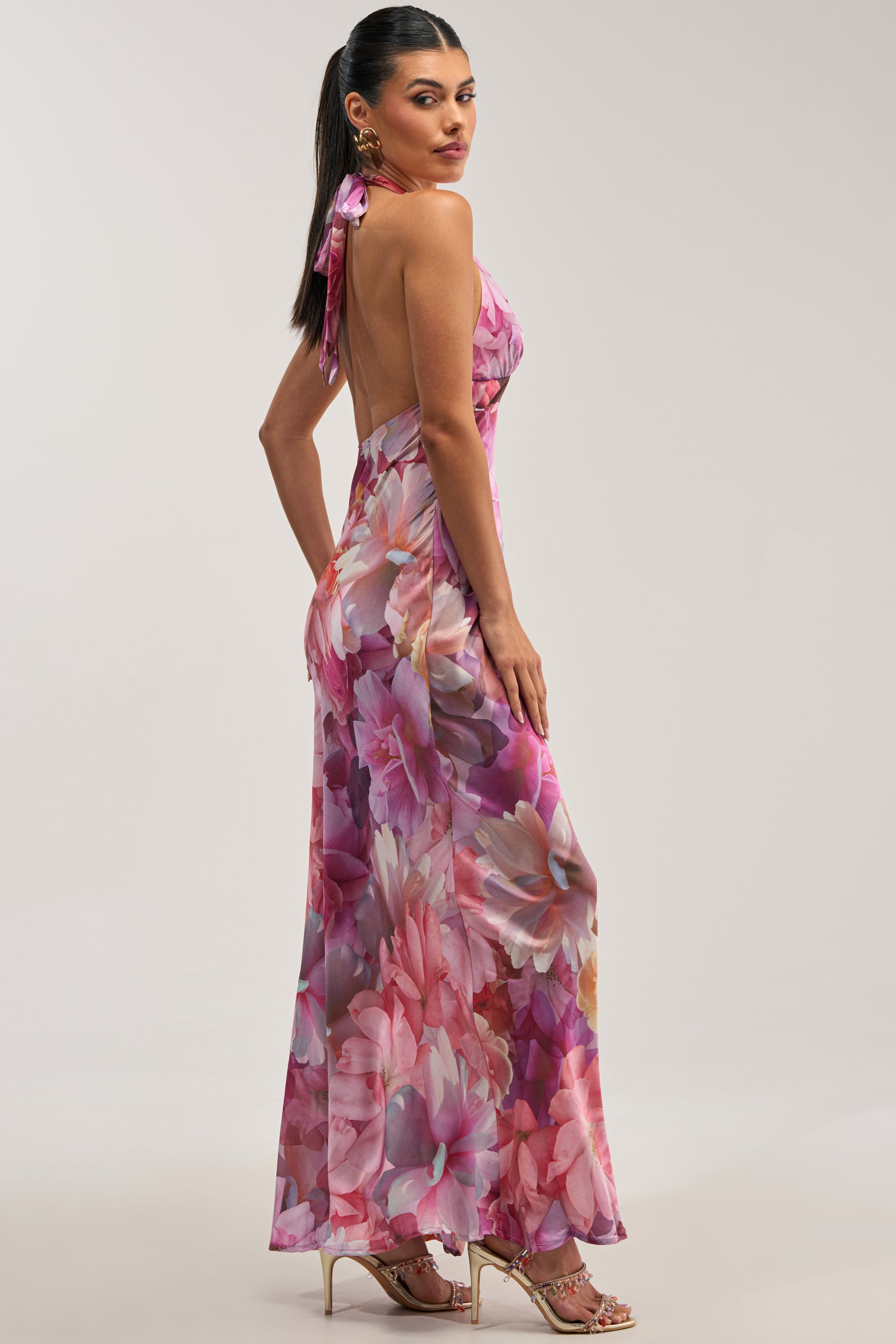 A woman models the HOPELESS ROMANTIC FLORAL SATIN MAXI DRESS, a pink halter maxi dress with an open back and flowing satin fabric. Adorned with large pink, purple, and peach blooms for a romantic flair, it’s styled here with gold strappy heels—perfect as a wedding guest ensemble.