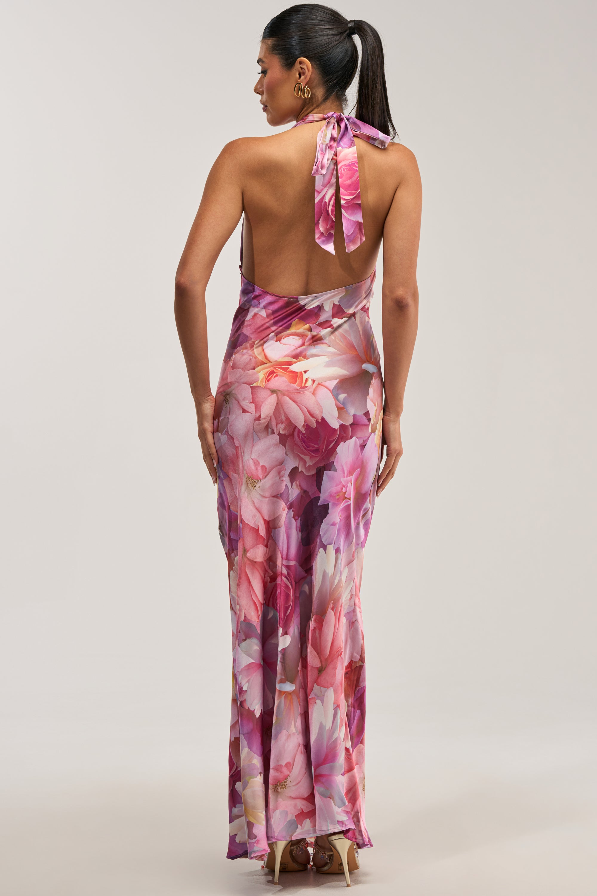A woman with a high ponytail and gold earrings models the HOPELESS ROMANTIC FLORAL SATIN MAXI DRESS—a floor-length, backless halter style showcasing a vibrant pink and purple floral pattern—as she poses against a light gray backdrop.