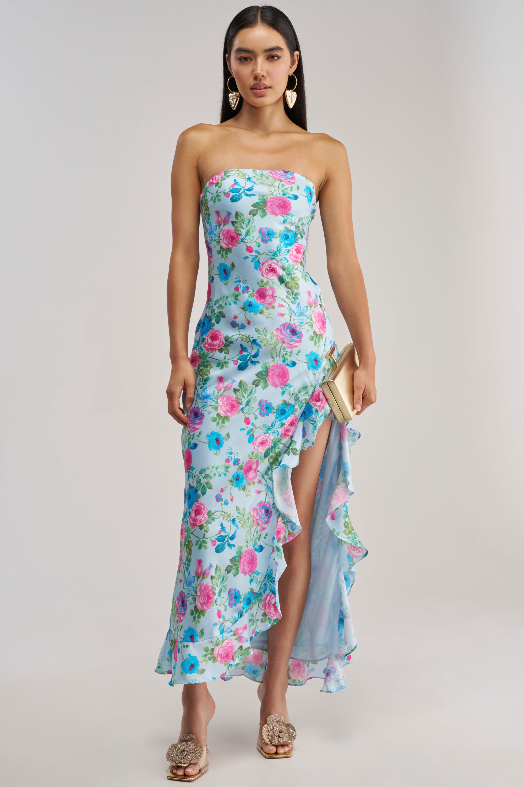A model showcases the SHE IS THE MOMENT FLORAL MAXI DRESS, a strapless maxi dress in light blue floral satin with a ruffled high slit. She accessorizes with gold statement earrings, a gold clutch, and embellished gold heels—an elegant ensemble ideal for a wedding guest. The shoot is set against a solid, light grey studio backdrop.