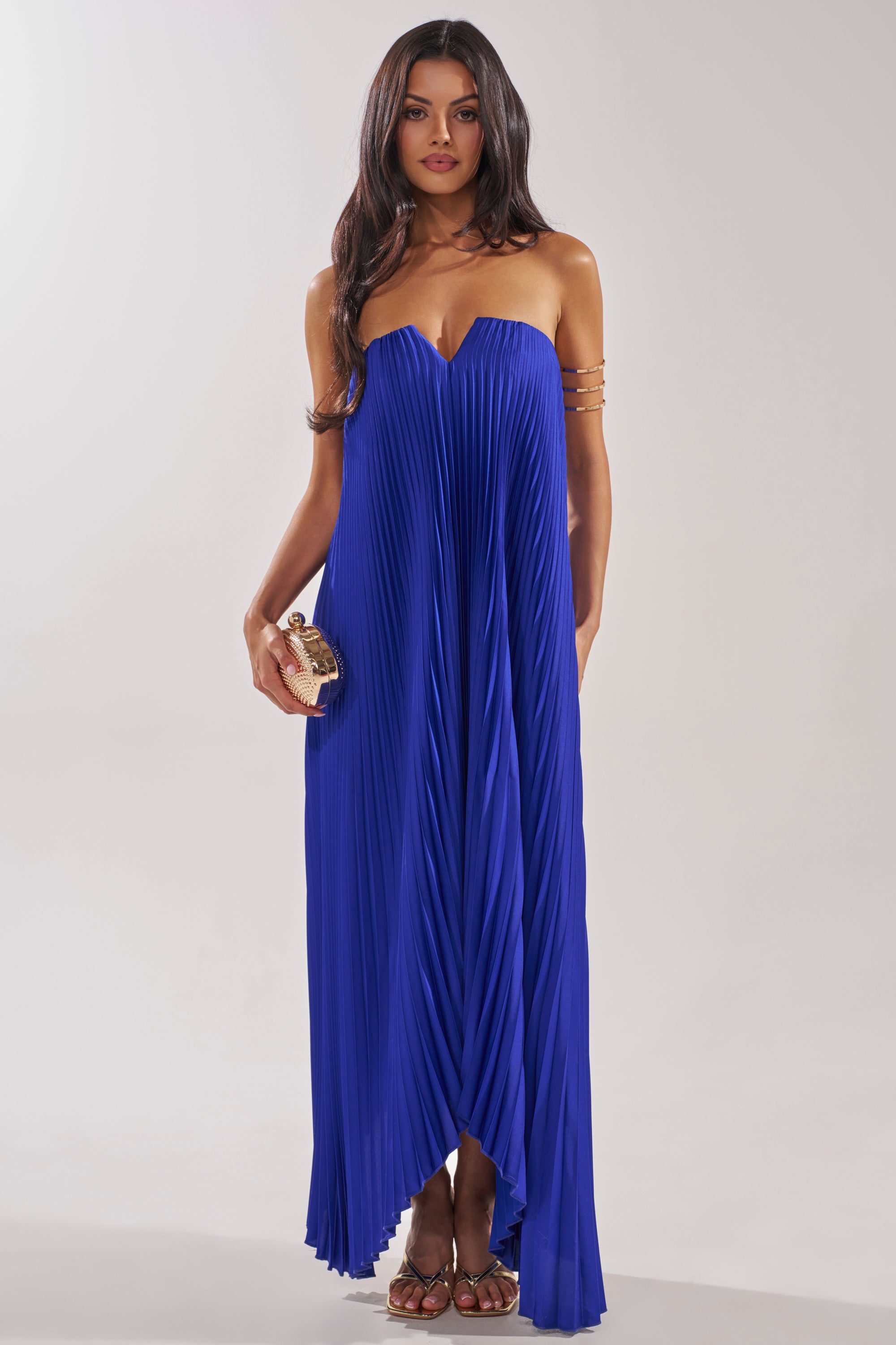 A woman models the EMERALD DREAM SLEEVELESS MAXI DRESS IN BLUE—strapless, pleated, with a sweetheart neckline—styled with a small clutch and arm cuffs, standing against a plain light background. 0
