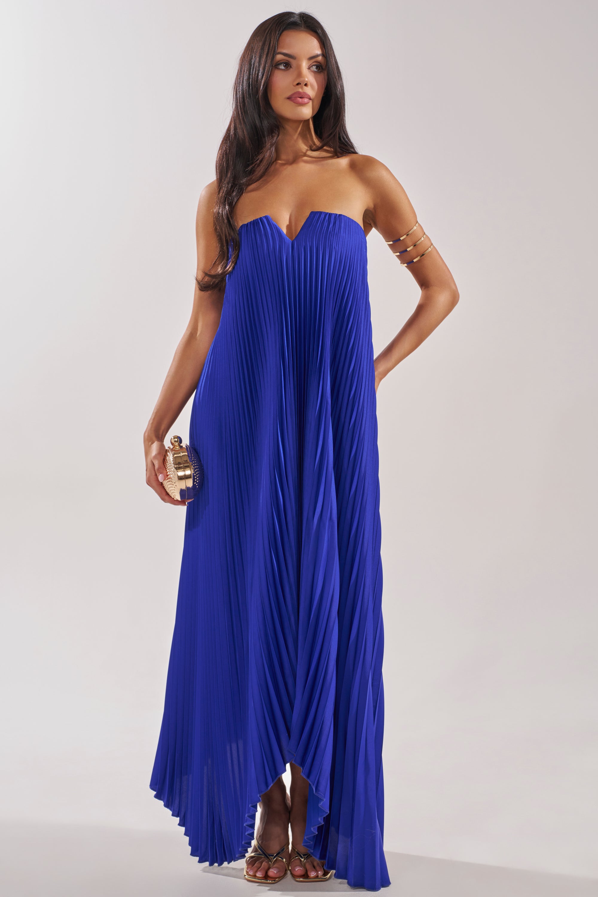 A woman in the EMERALD DREAM SLEEVELESS MAXI DRESS IN BLUE stands confidently, holding a gold clutch. She accessorizes with gold arm jewelry and sandals, her long dark hair flowing against a neutral background. 1