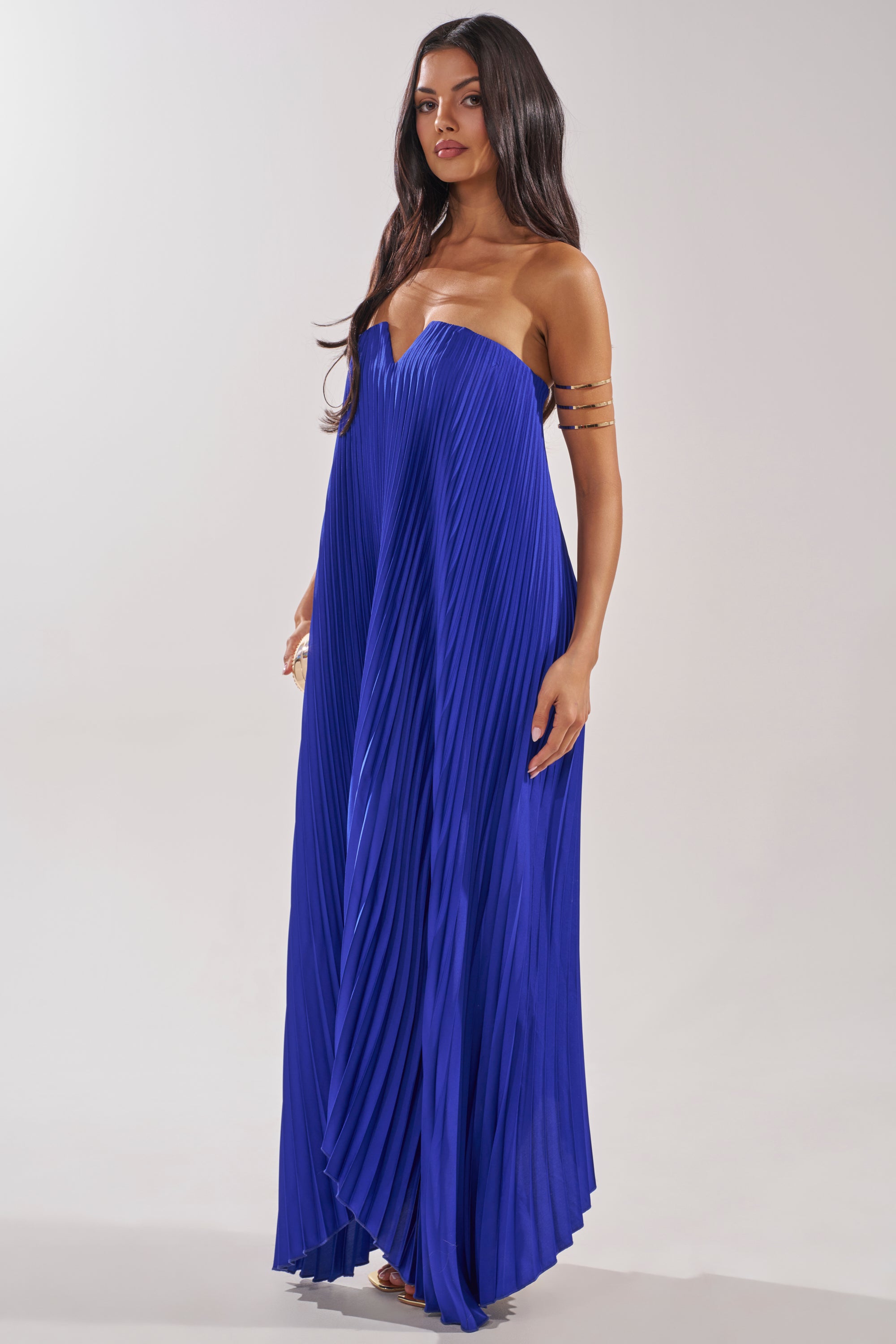 A woman with long dark hair wears the EMERALD DREAM SLEEVELESS MAXI DRESS IN BLUE with pleats and gold arm cuffs, standing against a plain light background. 2