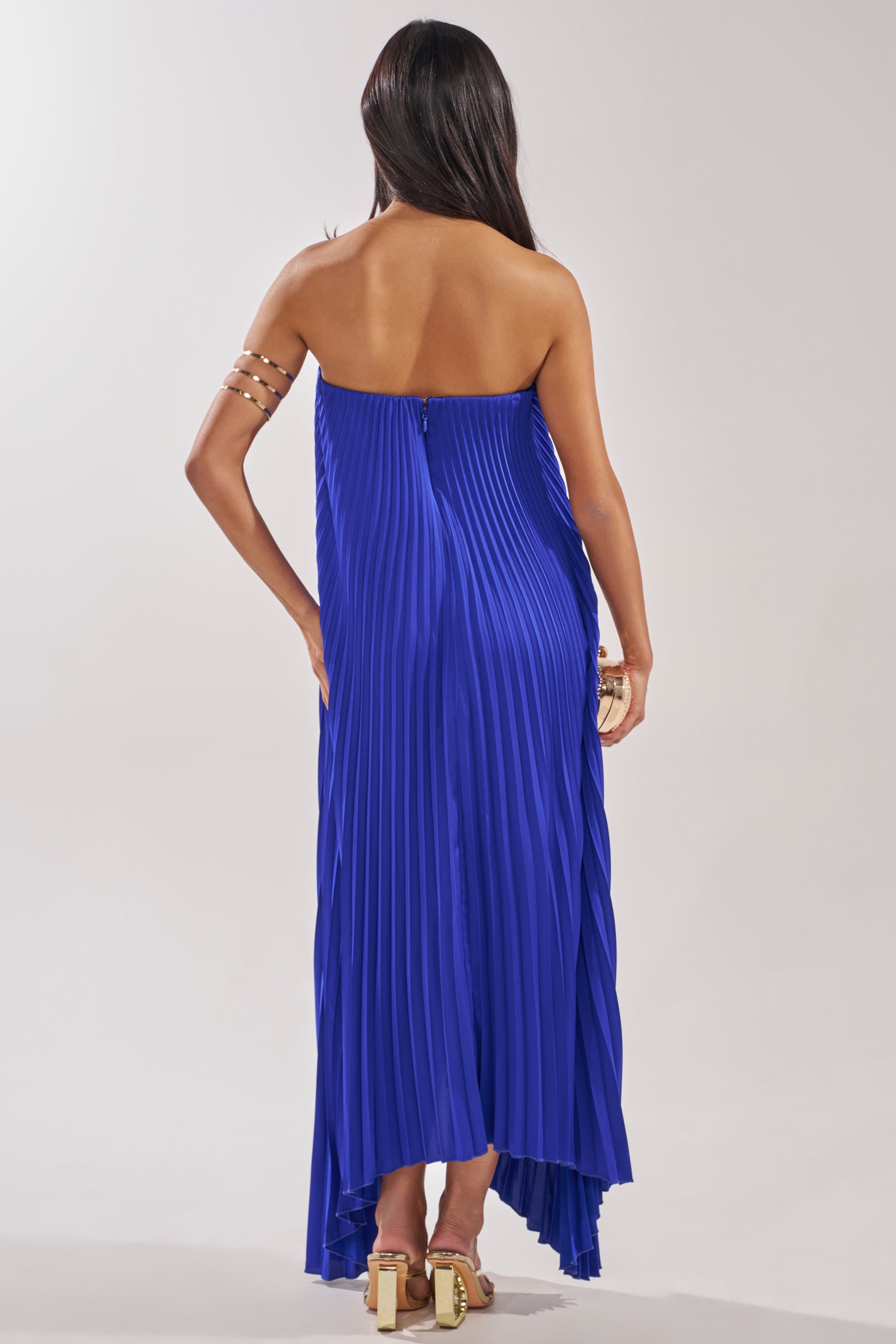 A woman faces away, wearing the EMERALD DREAM SLEEVELESS MAXI DRESS IN BLUE with pleats and an uneven hem, styled with gold arm cuffs and sandals. Her long dark hair cascades down her back against a neutral background. 3