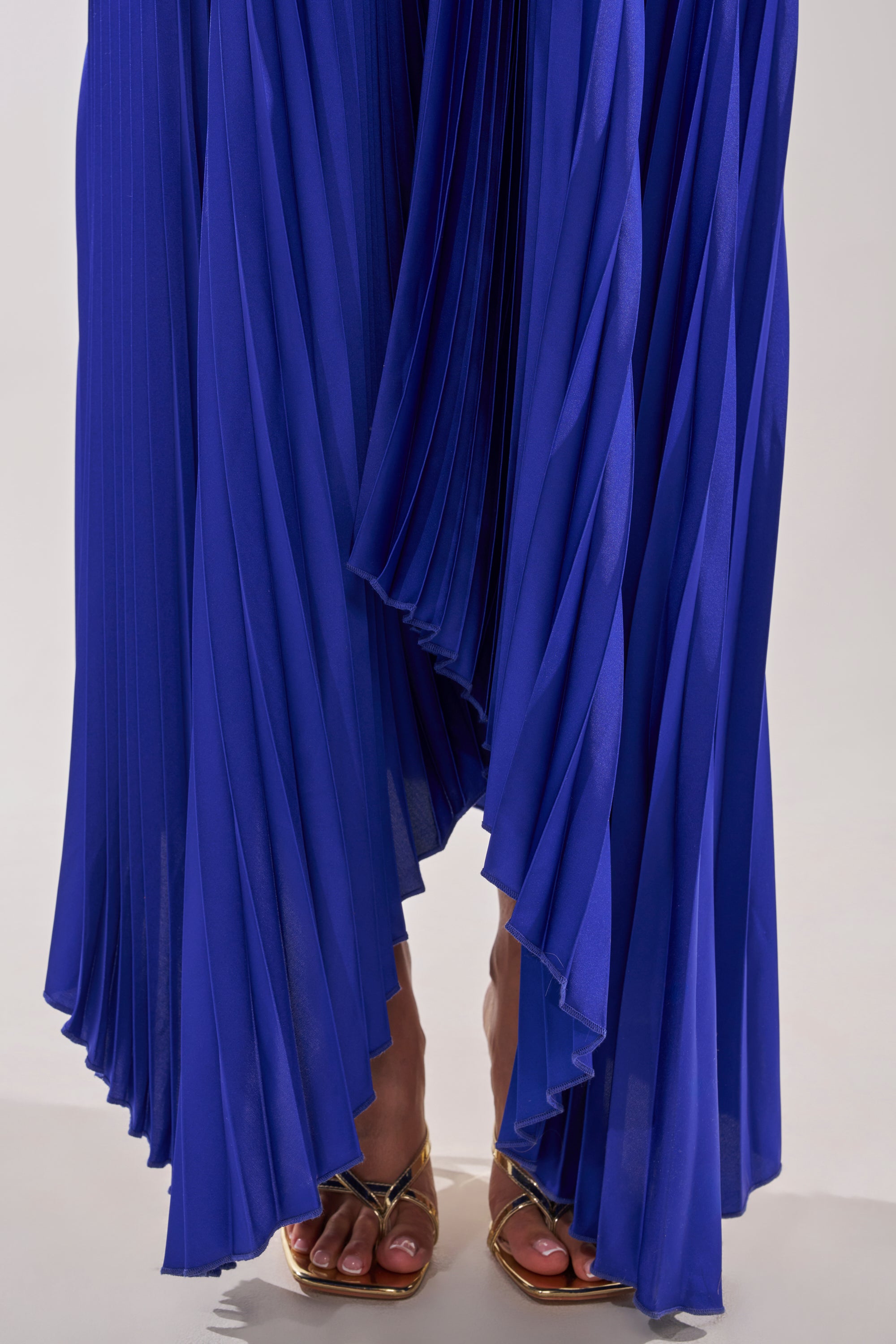 A person stands on a light surface wearing the EMERALD DREAM SLEEVELESS MAXI DRESS IN BLUE, a bright blue pleated evening dress that drapes to the ankles, paired with metallic open-toe heels. 5