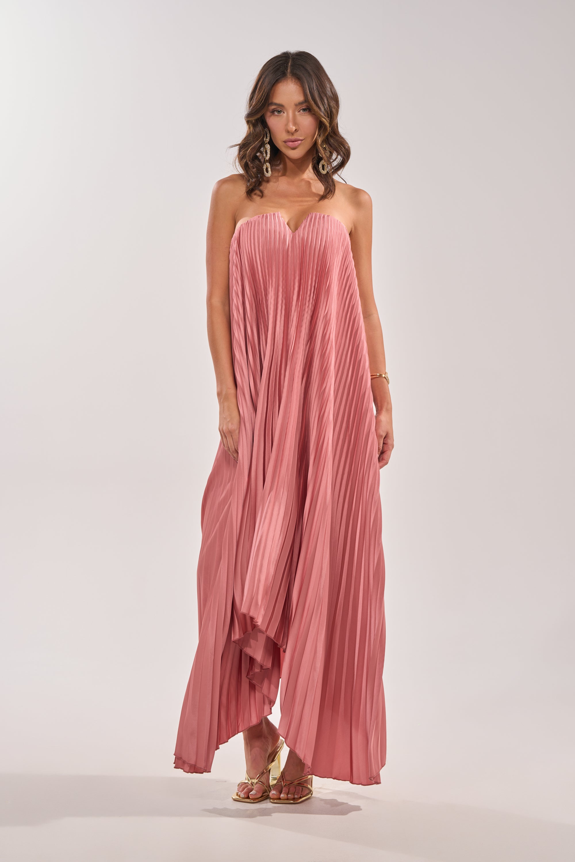 A woman stands against a plain background wearing the EMERALD DREAM SLEEVELESS MAXI DRESS IN PINK, a flowing pleated formal gown. She has wavy hair, gold earrings, and sandals, with her arms relaxed at her sides. 3