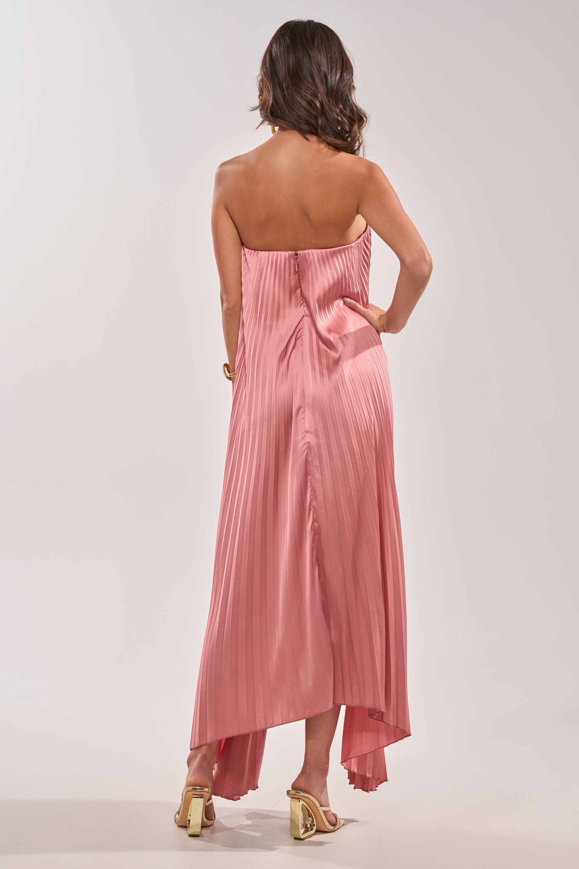 A woman with wavy brown hair, wearing the EMERALD DREAM SLEEVELESS MAXI DRESS IN PINK and beige heeled sandals, stands facing away from the camera against a plain light background. 5