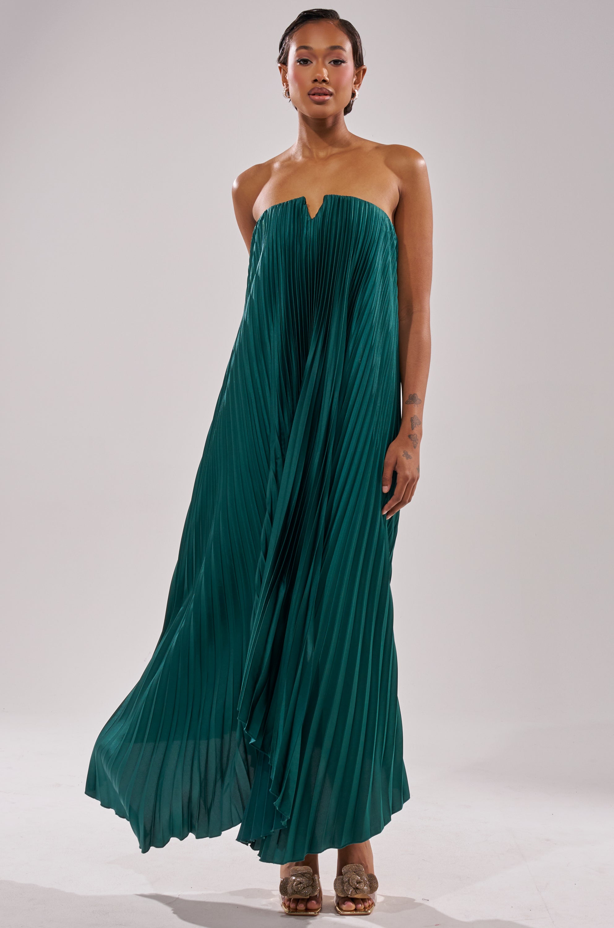 A person models the EMERALD DREAM SLEEVELESS MAXI DRESS in emerald green satin with pleats, paired with gold open-toed heels, against a plain light background.