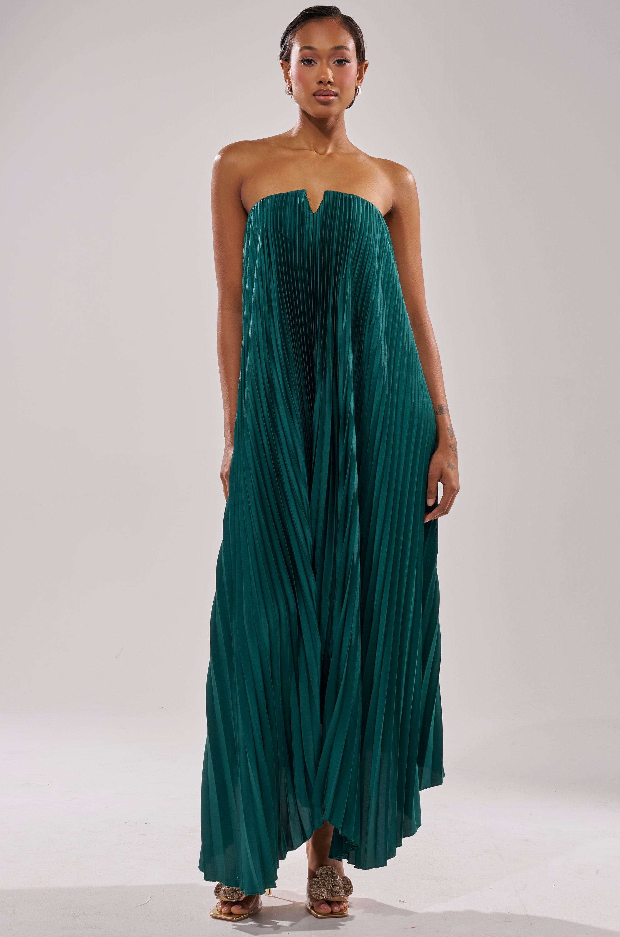 A woman models the EMERALD DREAM SLEEVELESS MAXI DRESS, featuring pleats and a notched strapless neckline. She has short hair and poses with a neutral expression against a plain background.