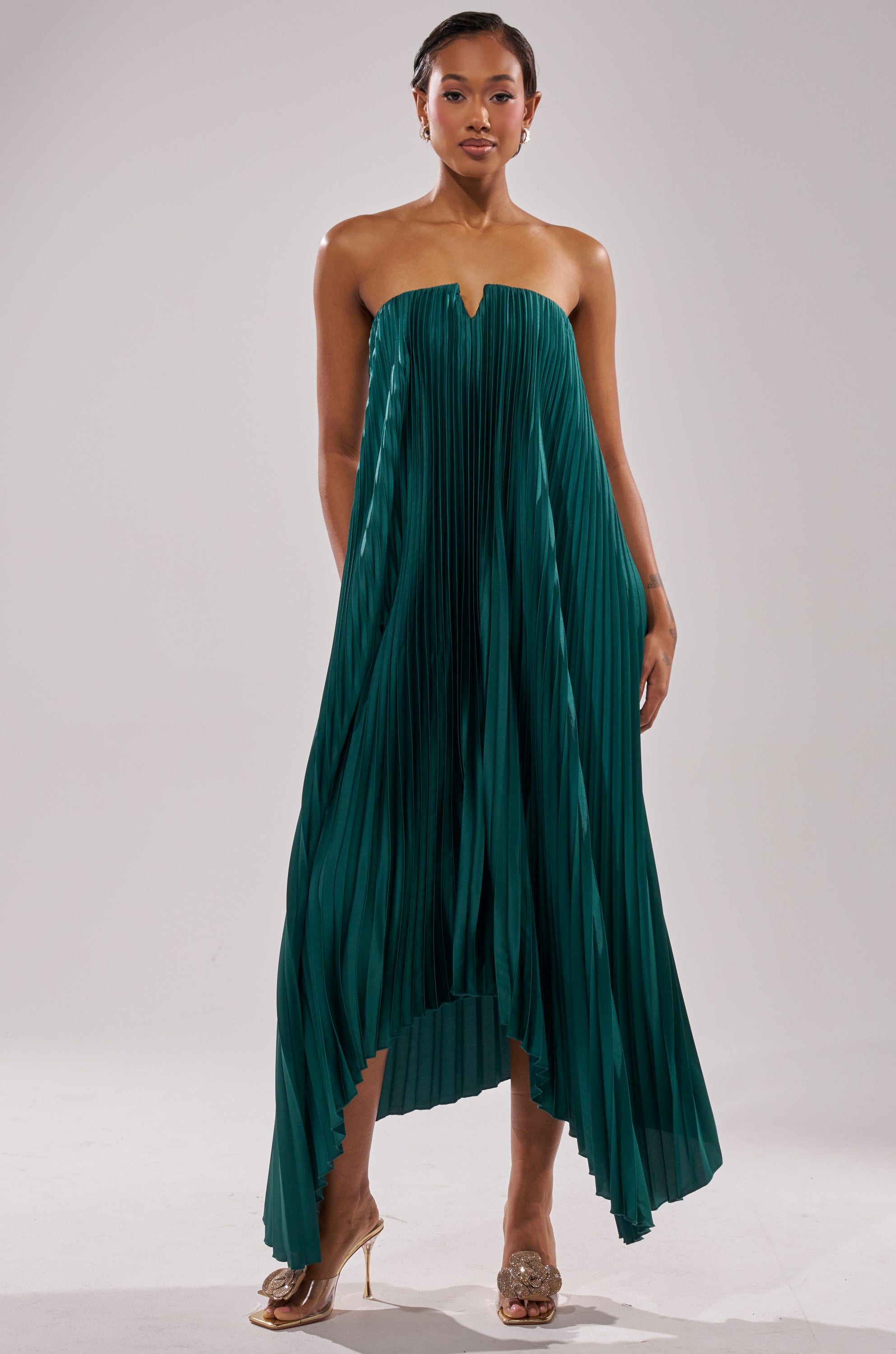 A woman with short hair models the EMERALD DREAM SLEEVELESS MAXI DRESS, a pleated emerald green maxi with an asymmetrical hemline. She pairs it with gold open-toe heels and poses against a plain light background.
