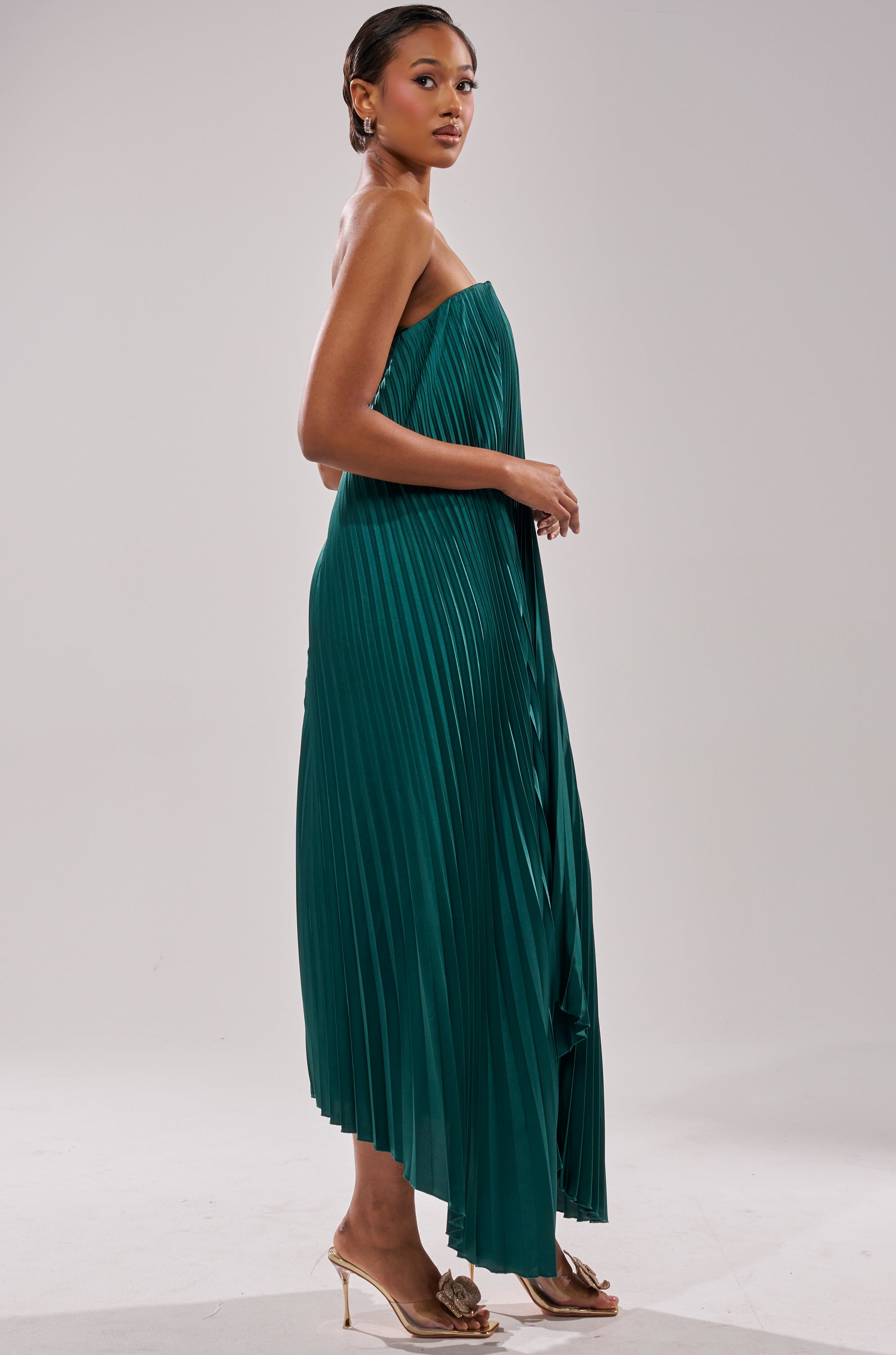 A woman stands sideways in the EMERALD DREAM SLEEVELESS MAXI DRESS—an emerald green pleated maxi. She pairs it with gold heels, hair pulled back, and poses against a plain light background.