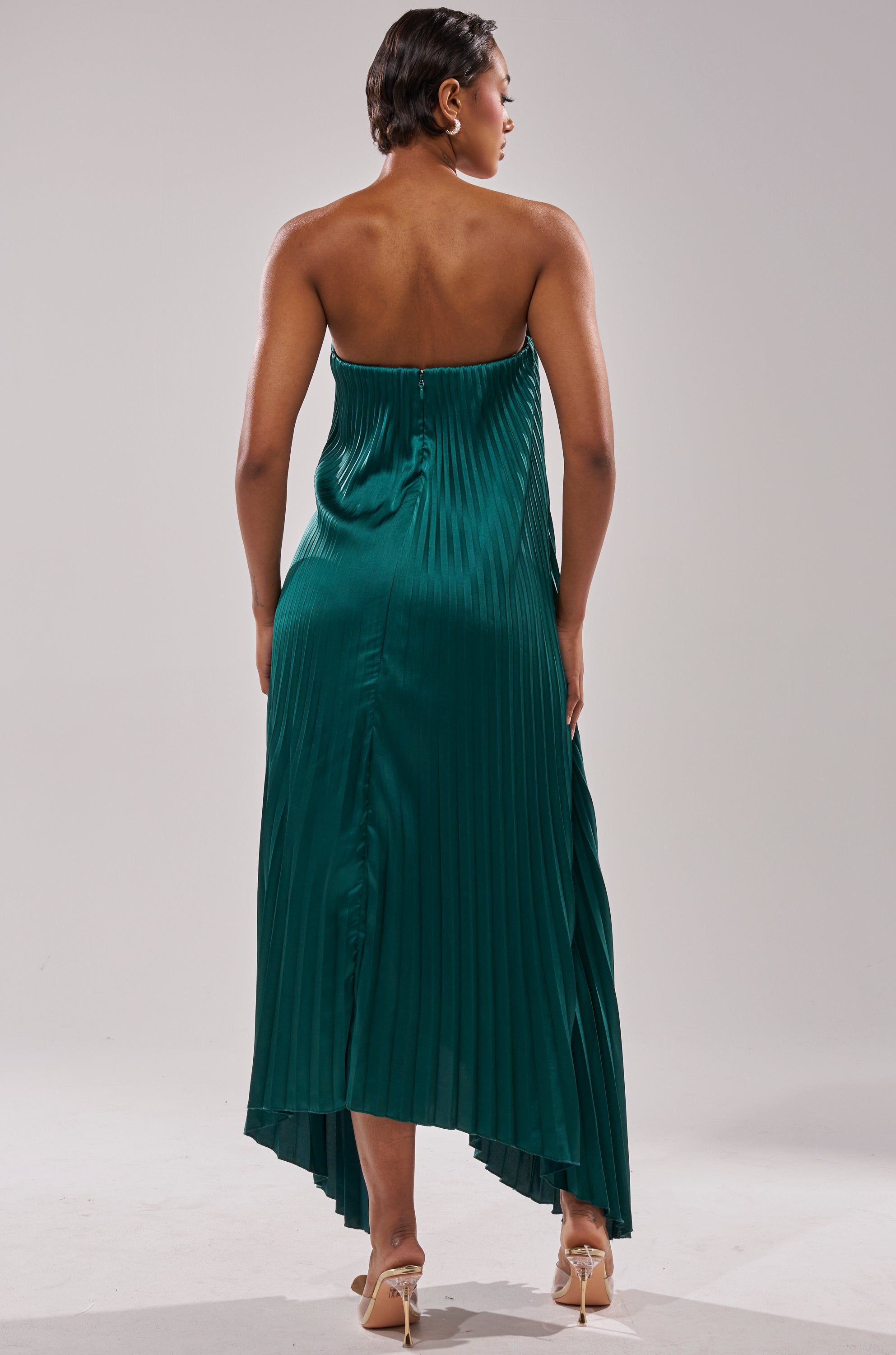 A woman with short hair models the EMERALD DREAM SLEEVELESS MAXI DRESS, featuring a pleated strapless design and asymmetrical hem in emerald green. She pairs it with nude heels, posing against a plain light background.