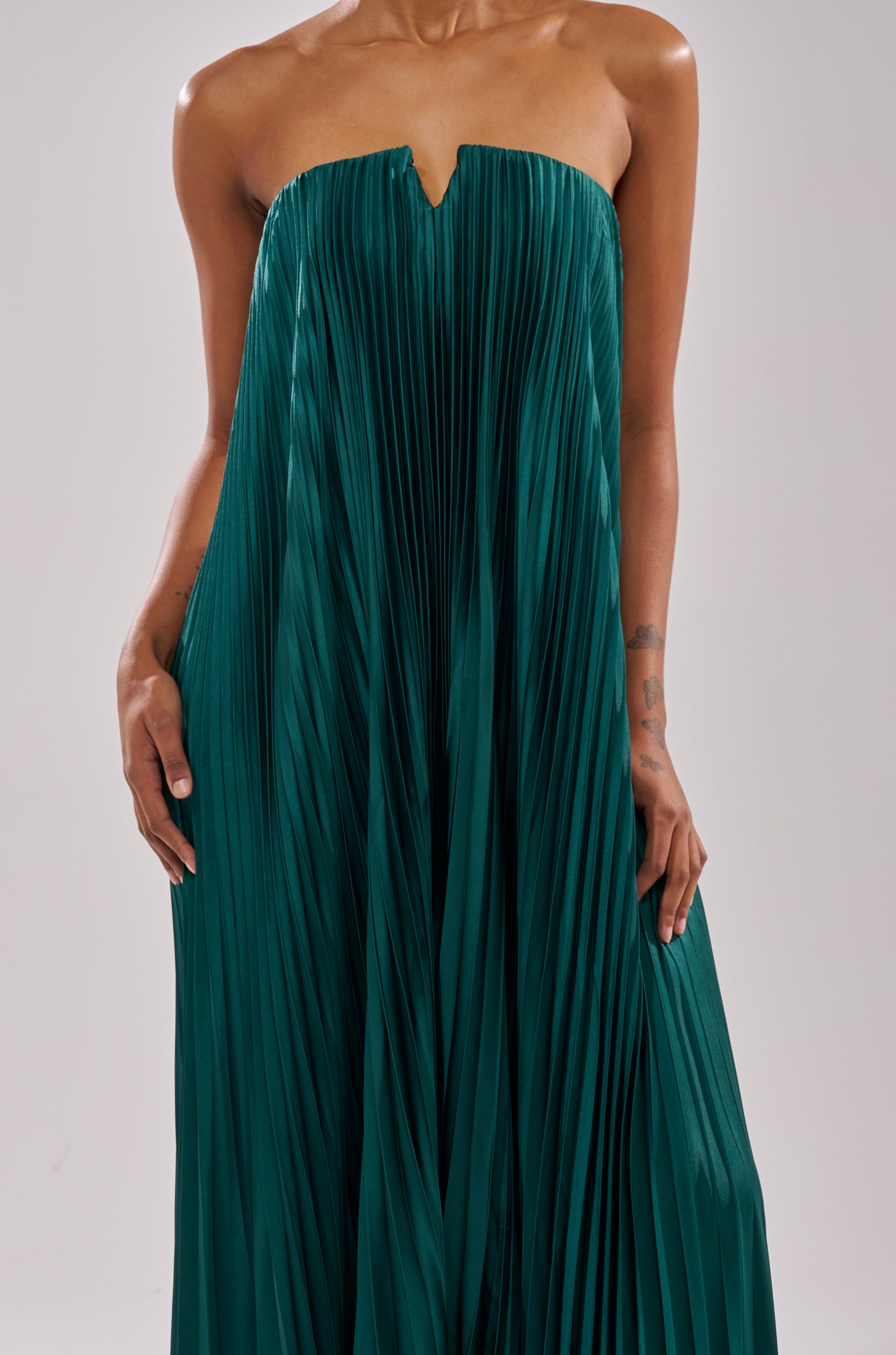Someone stands against a plain light background wearing the EMERALD DREAM SLEEVELESS MAXI DRESS, a pleated deep green gown with a structured neckline. Their arms are relaxed at their sides.