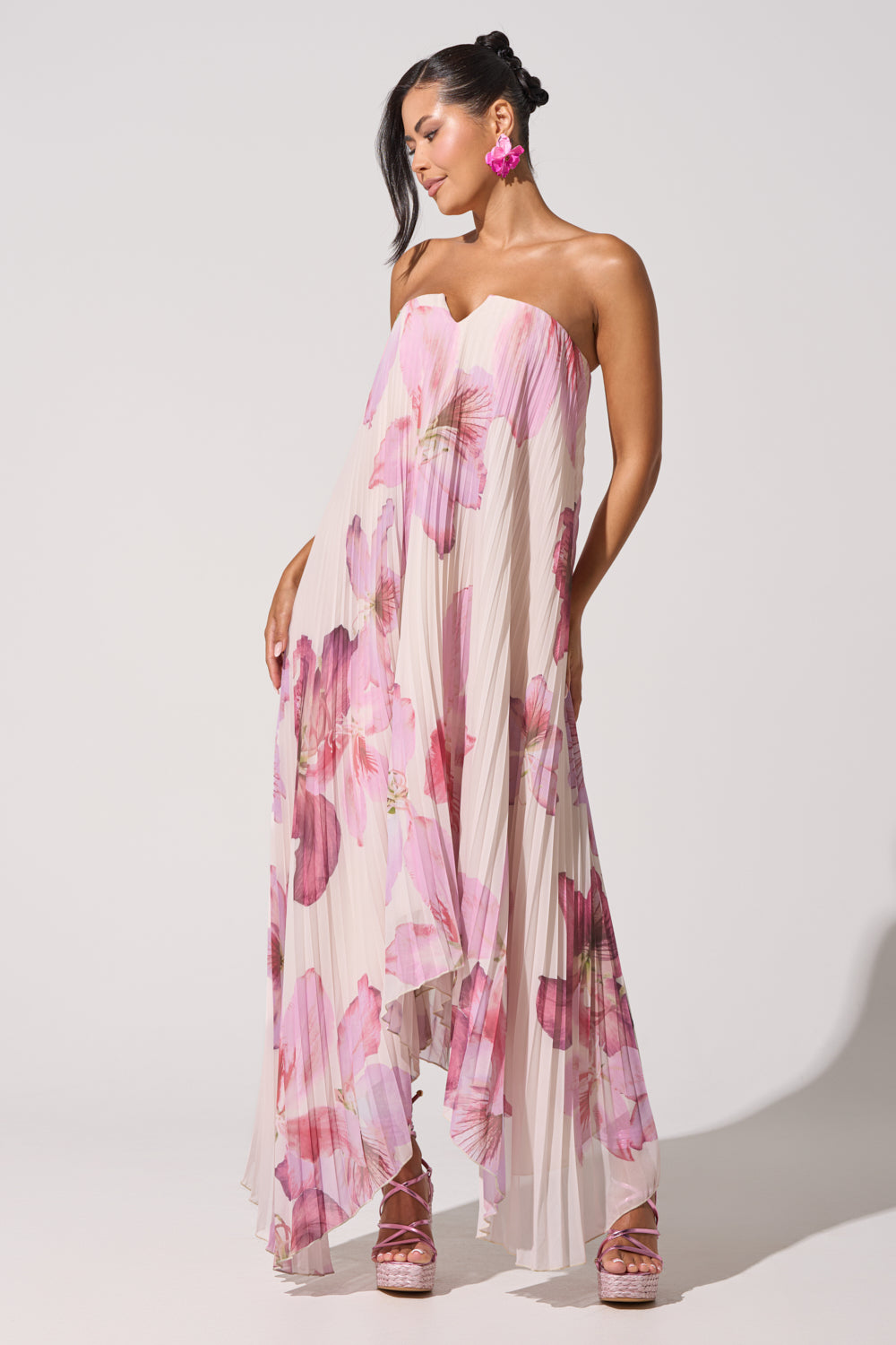 A woman models the JULIANA STRAPLESS FLORAL MAXI DRESS, featuring pink and purple pleats. She completes the look with large pink earrings, matching platform heels, and her dark hair styled to one side. 2