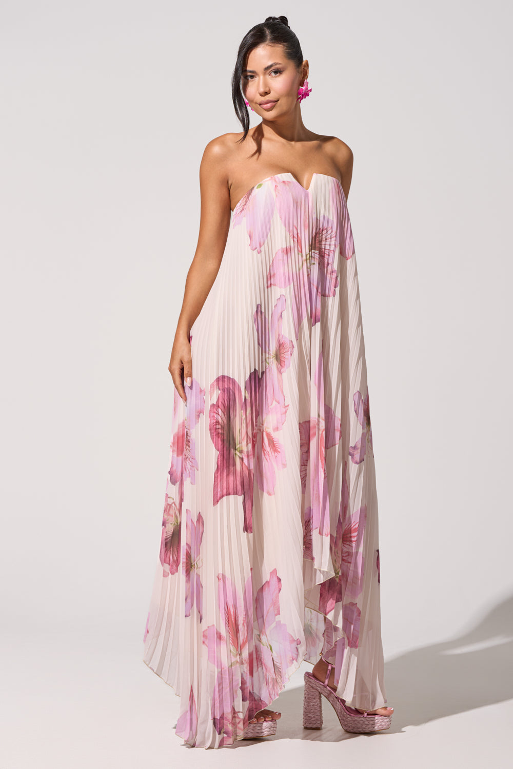 A woman models the JULIANA STRAPLESS FLORAL MAXI DRESS in white with bold pink floral prints, styled with pink heels and flower earrings against a plain white background for a chic formal summer look. 11