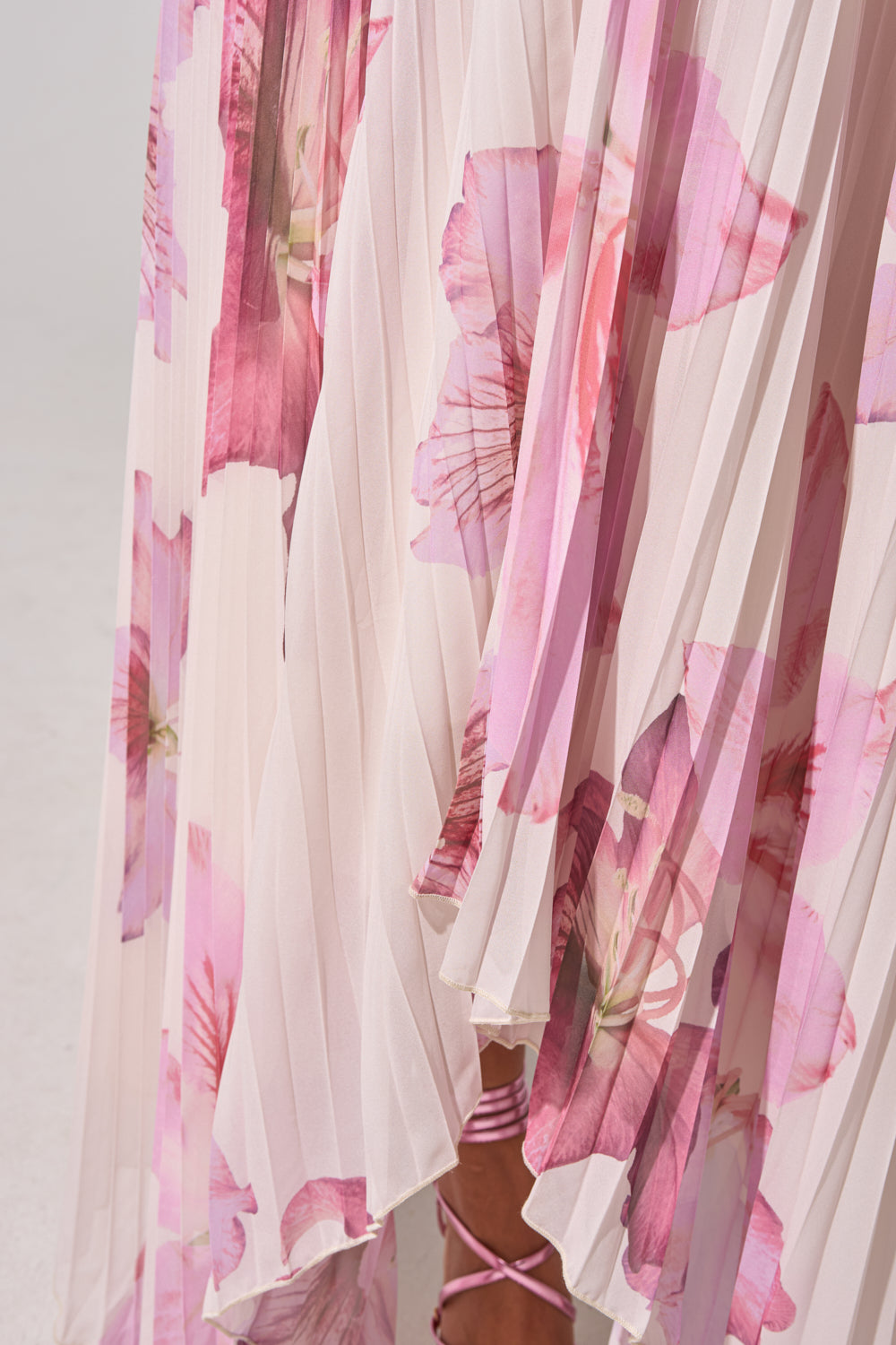 Close-up of the JULIANA STRAPLESS FLORAL MAXI DRESS, featuring pink pleats and bold floral prints. A peek of leg with pink lace-up sandals emerges from beneath the flowing sheer fabric, perfect for a formal summer look. 12