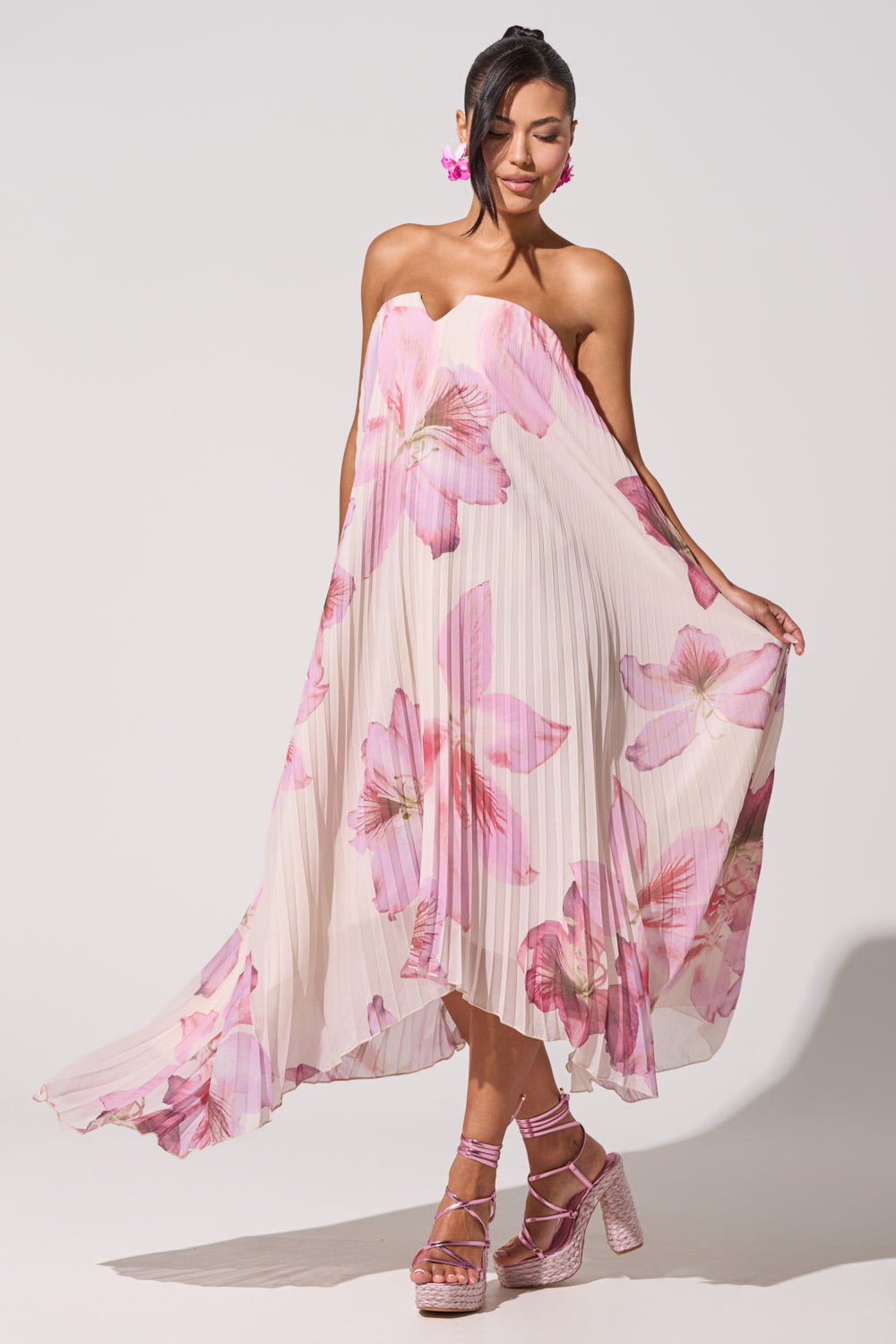Wearing the JULIANA STRAPLESS FLORAL MAXI DRESS, she holds the sides of her bold pink floral dress, pairing it with pink platform heels, floral earrings, and side-swept hair for a chic formal summer look. 3