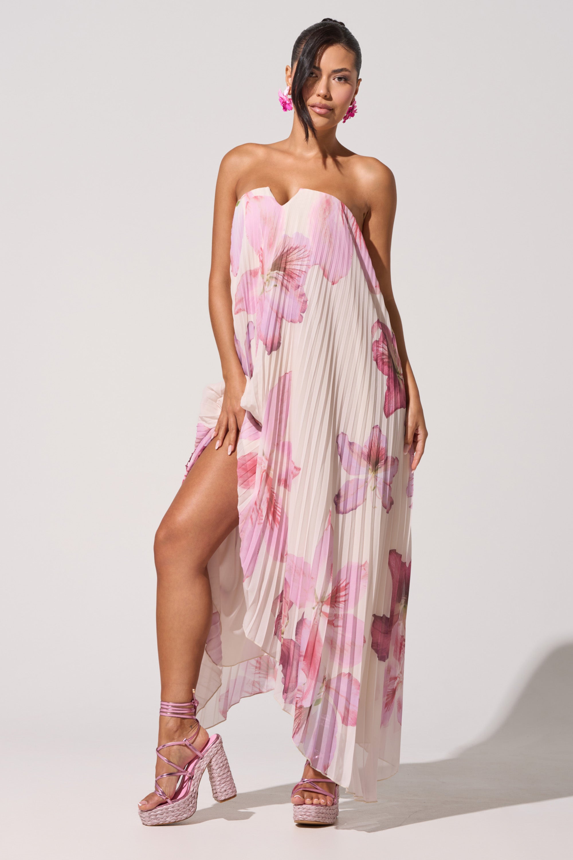 A woman models the JULIANA STRAPLESS FLORAL MAXI DRESS, featuring pleated floral prints and an asymmetrical hem, styled with pink platform heels and flower earrings, her hair swept to one side. 1