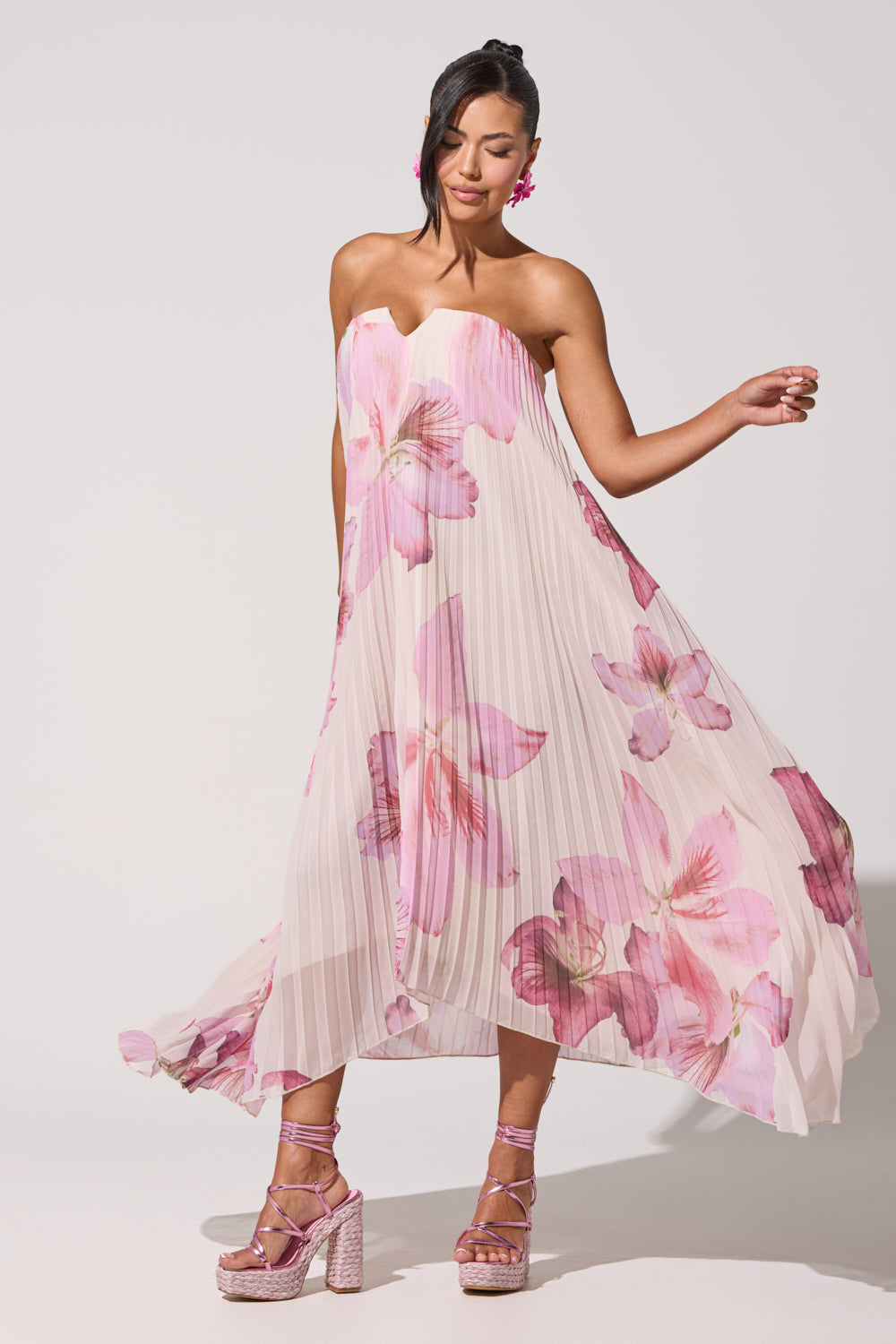 A woman poses in the JULIANA STRAPLESS FLORAL MAXI DRESS, a pink strapless pleated maxi with bold floral prints, styled with pink platform heels and flower earrings against a plain white background. 4