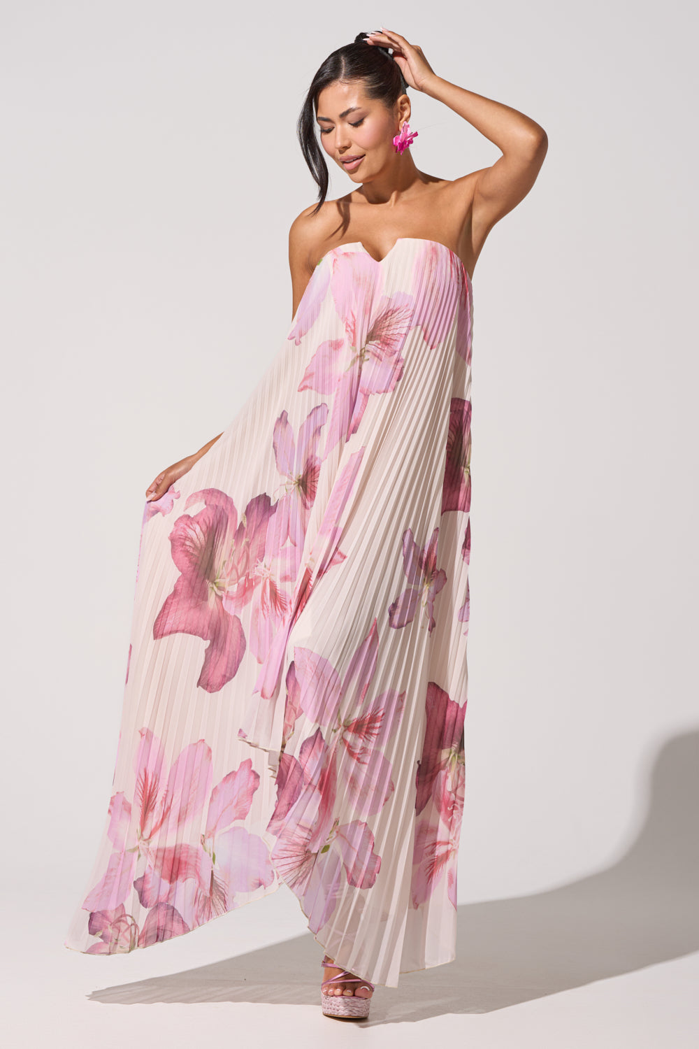 A woman poses in the JULIANA STRAPLESS FLORAL MAXI DRESS featuring large pink prints. She pairs it with pink flower earrings and platform sandals, touching her hair with one hand and holding the dress with the other against a plain white background. 5