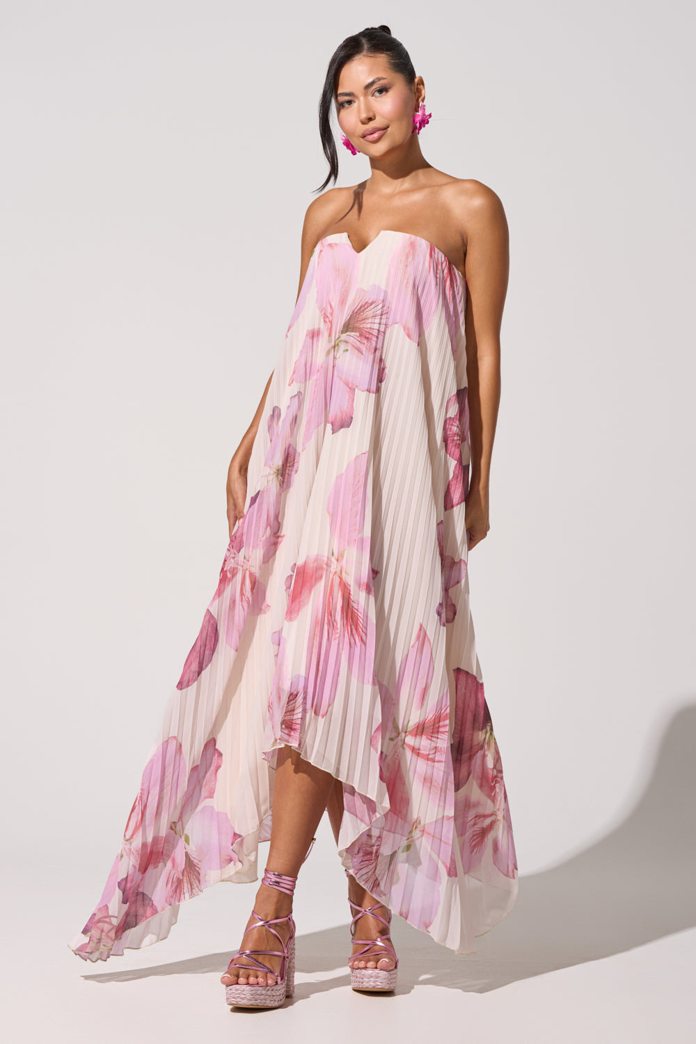 Wearing the JULIANA STRAPLESS FLORAL MAXI DRESS with a high-low hem and vivid pink blooms, she pairs it with pink platform sandals and floral earrings, standing against a light background. 6