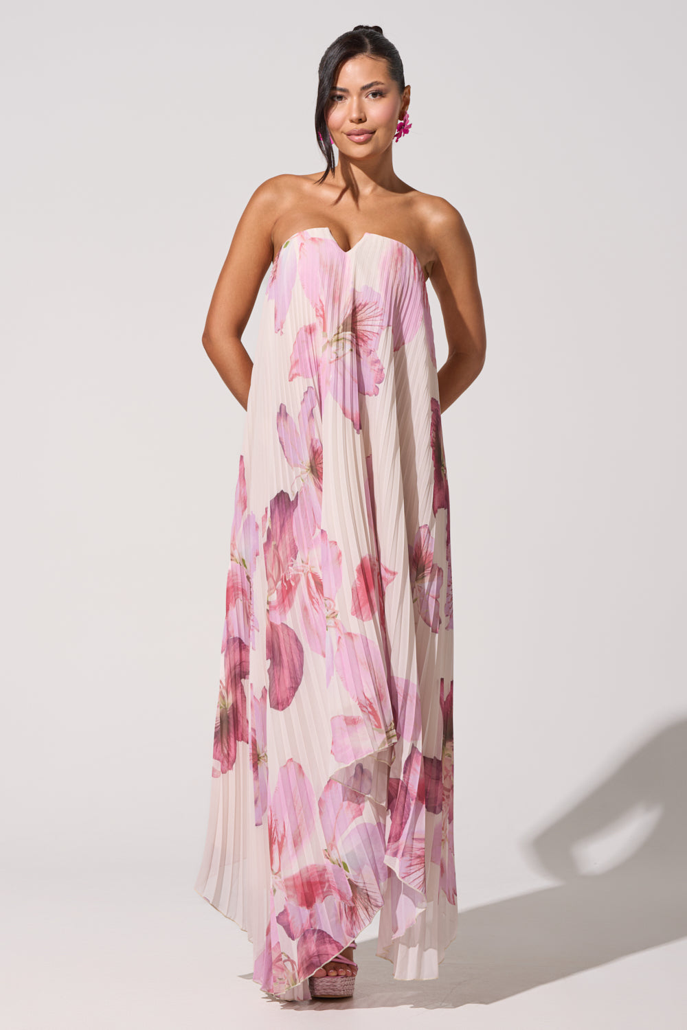 A woman stands against a plain background in the JULIANA STRAPLESS FLORAL MAXI DRESS with bold pink prints, smiling softly with her dark hair to one side—an elegant choice for formal summer occasions. 7