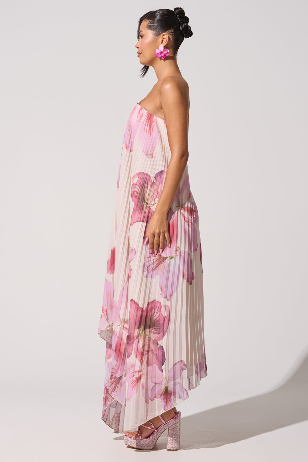 A woman in profile wears the JULIANA STRAPLESS FLORAL MAXI DRESS, featuring bold pink blossoms. She accessorizes with pink flower earrings and platform heels, her hair styled in an elegant updo against a plain background. 8
