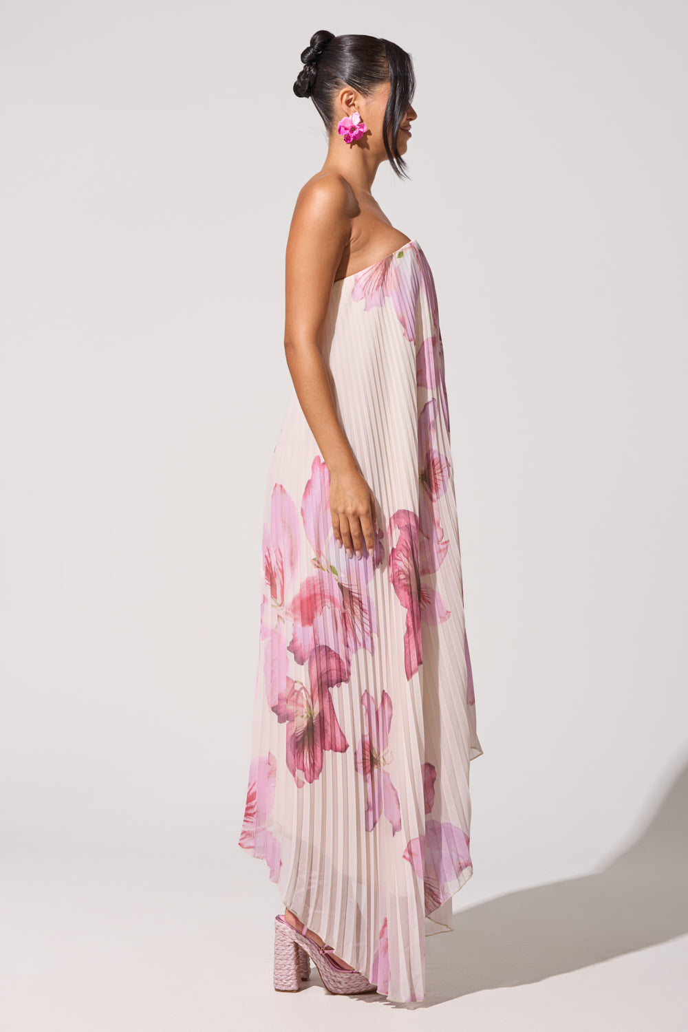 A woman in the JULIANA STRAPLESS FLORAL MAXI DRESS, featuring pleats, stands in profile. Her hair is in an updo, and she accessorizes with pink statement earrings and matching pink platform shoes against a plain light background. 9