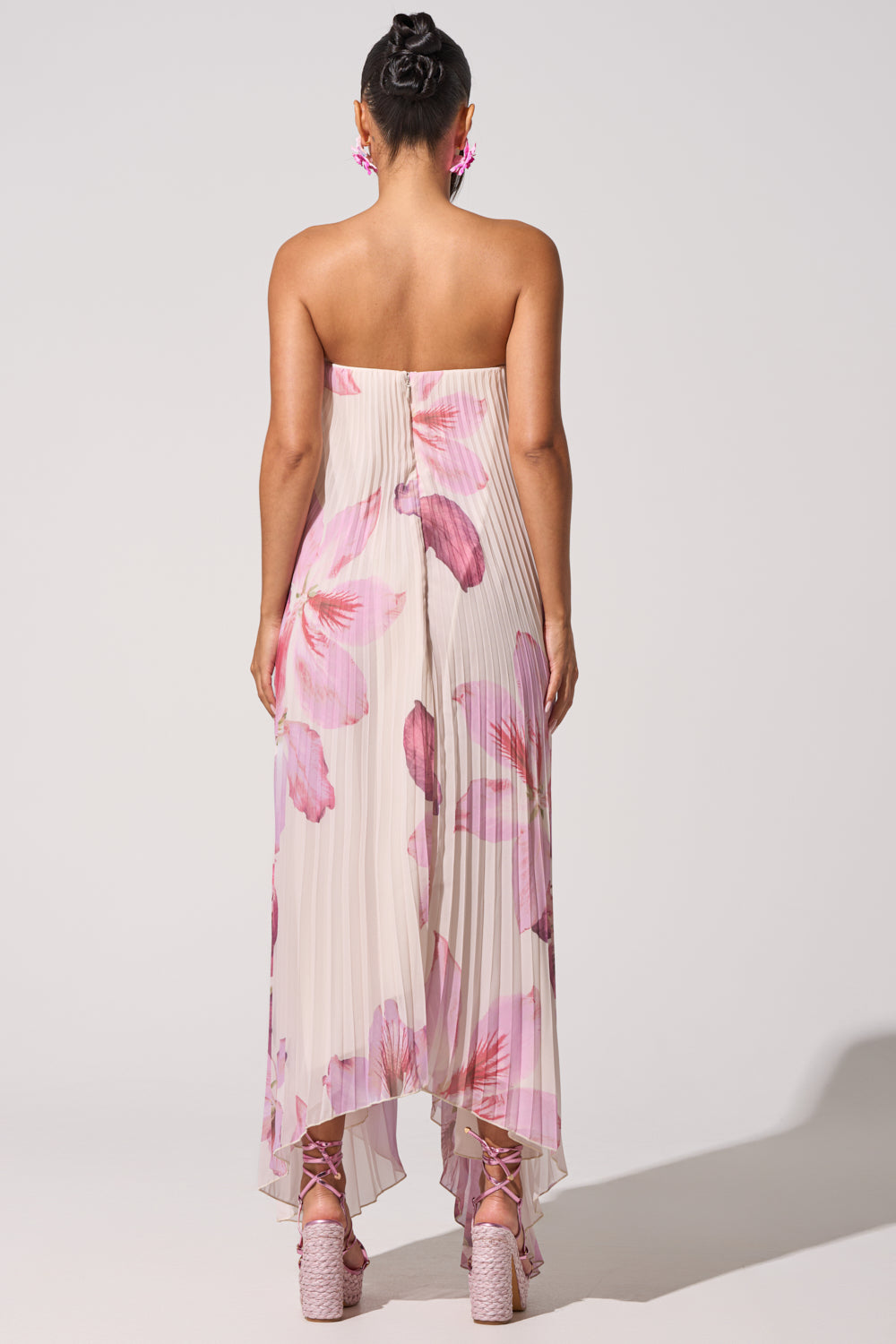 A woman faces away, wearing the JULIANA STRAPLESS FLORAL MAXI DRESS—pink with large floral prints. Her hair is in a bun, and she pairs pink earrings with chunky platform sandals against a simple, light backdrop. 10