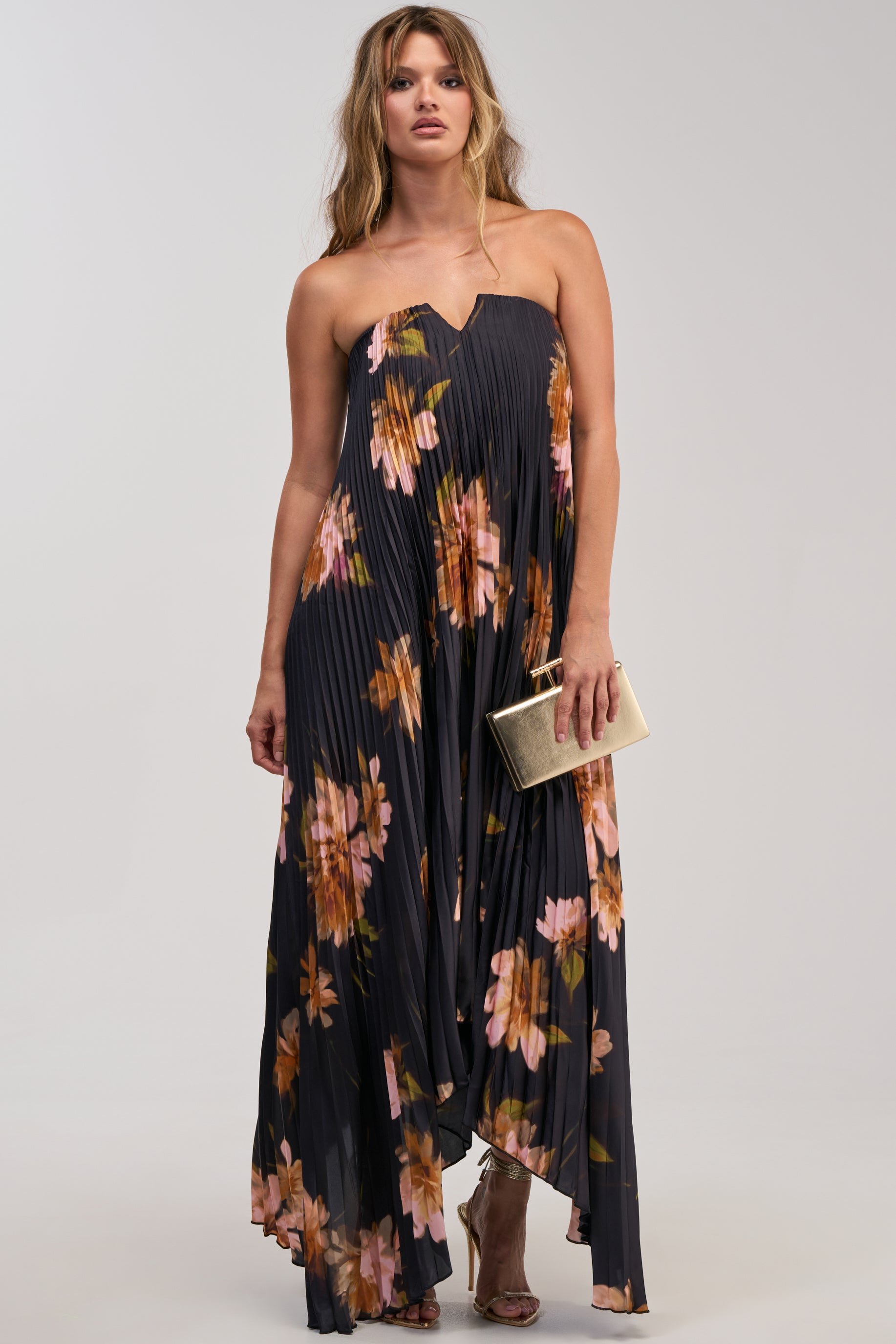 Woman wearing the MEET ME IN ITALY SATIN MIDI DRESS IN BLACK MULTI, a strapless black dress featuring a pink and orange floral print and pleated silhouette, styled with a gold clutch and strappy heels, standing against a plain background.
