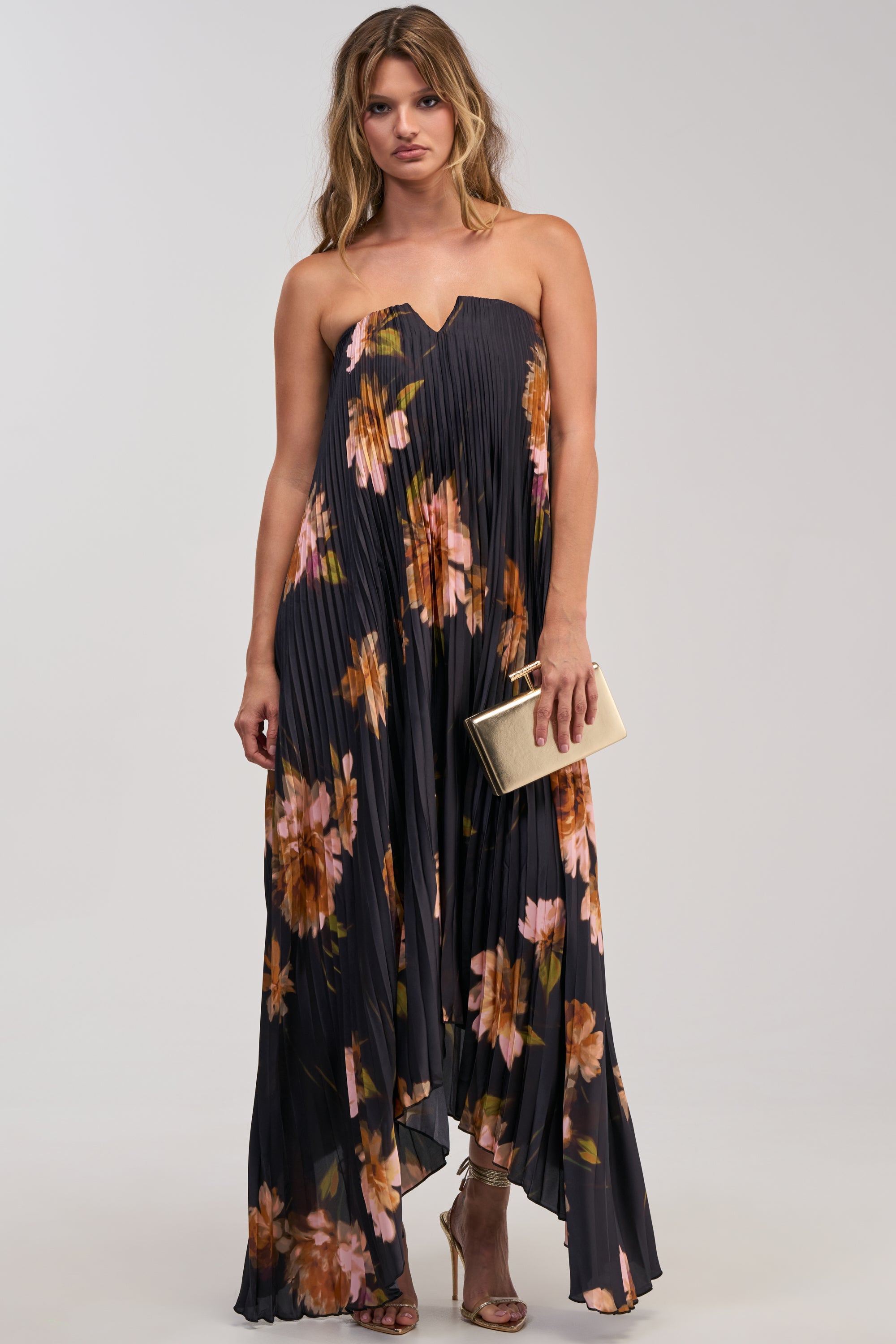 Wearing the MEET ME IN ITALY SATIN MIDI DRESS IN BLACK MULTI—a black, strapless floral midi dress adorned with vibrant pink and orange blooms—a woman completes her effortlessly chic look with a gold clutch and strappy gold heels, standing against a plain light background for the perfect formal outfit.