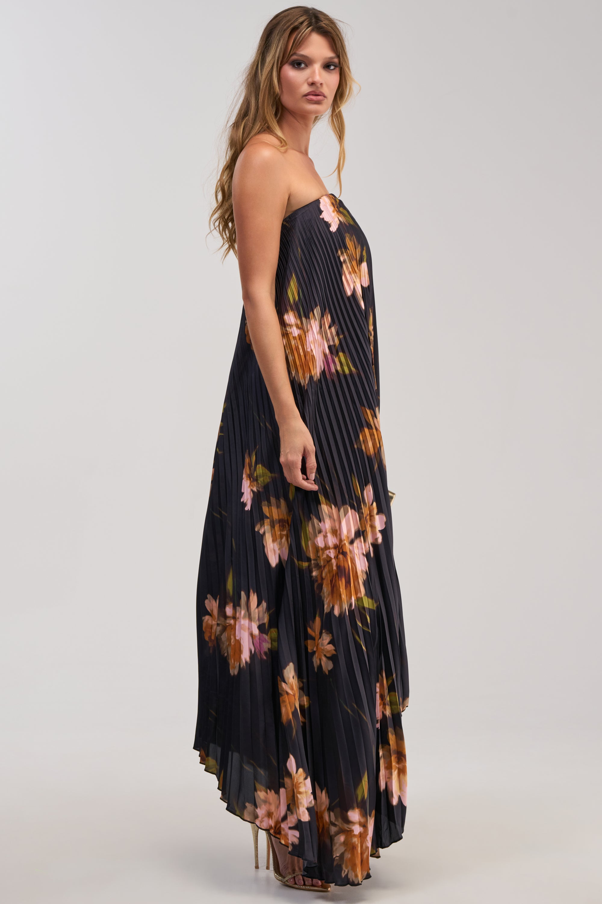 Wearing the MEET ME IN ITALY SATIN MIDI DRESS IN BLACK MULTI, a woman stands sideways against a plain light background. This elegant dress, featuring a black base with vibrant pink and orange floral prints and pleated details, is perfect for formal occasions—highlighting sophistication and grace.