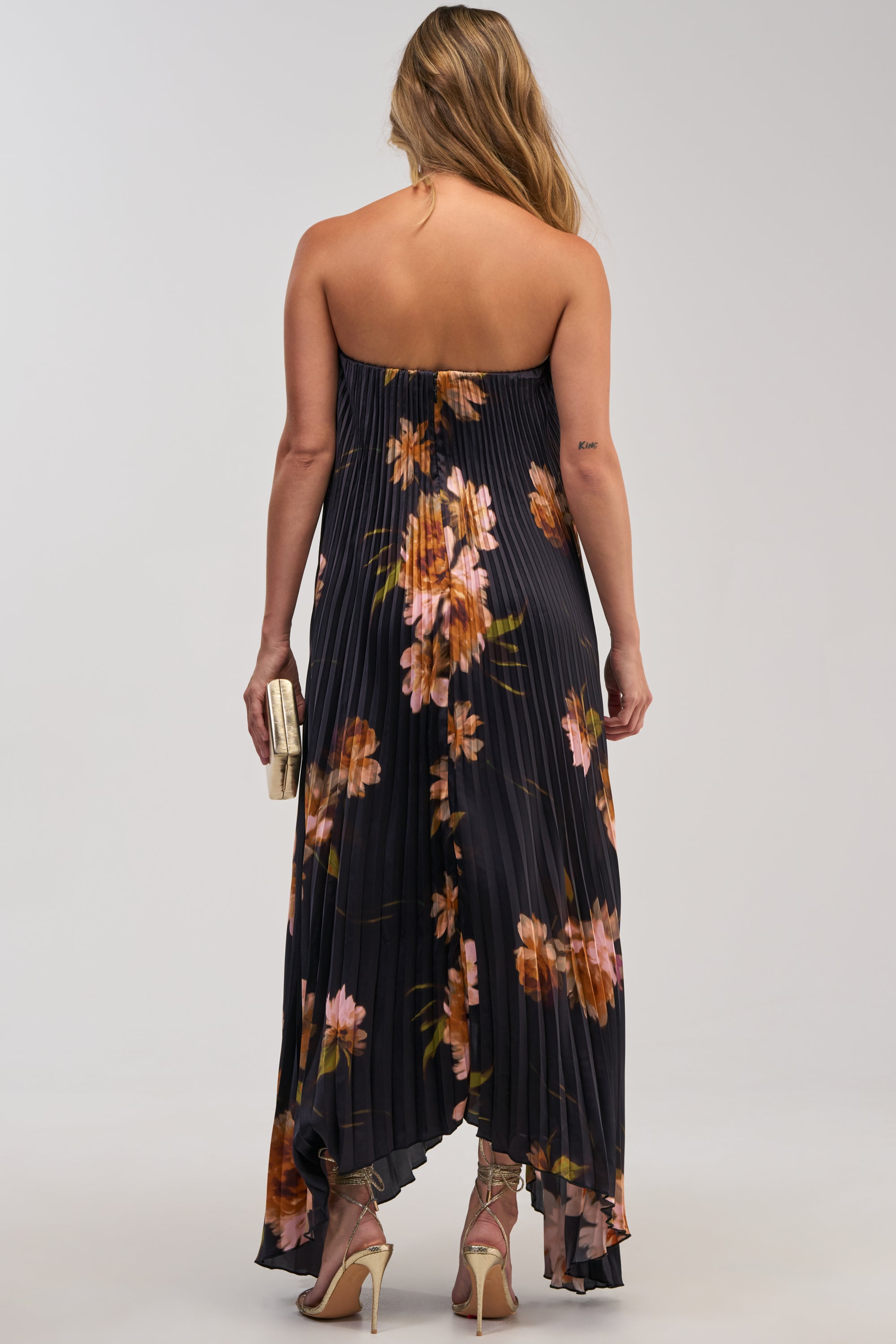 A woman with long hair wears the MEET ME IN ITALY SATIN MIDI DRESS IN BLACK MULTI, a strapless dress featuring pleated, dark satin fabric adorned with pink and orange patterns. Styled for a formal occasion, she holds a clutch and stands in high heels, facing away from the camera against a plain background.
