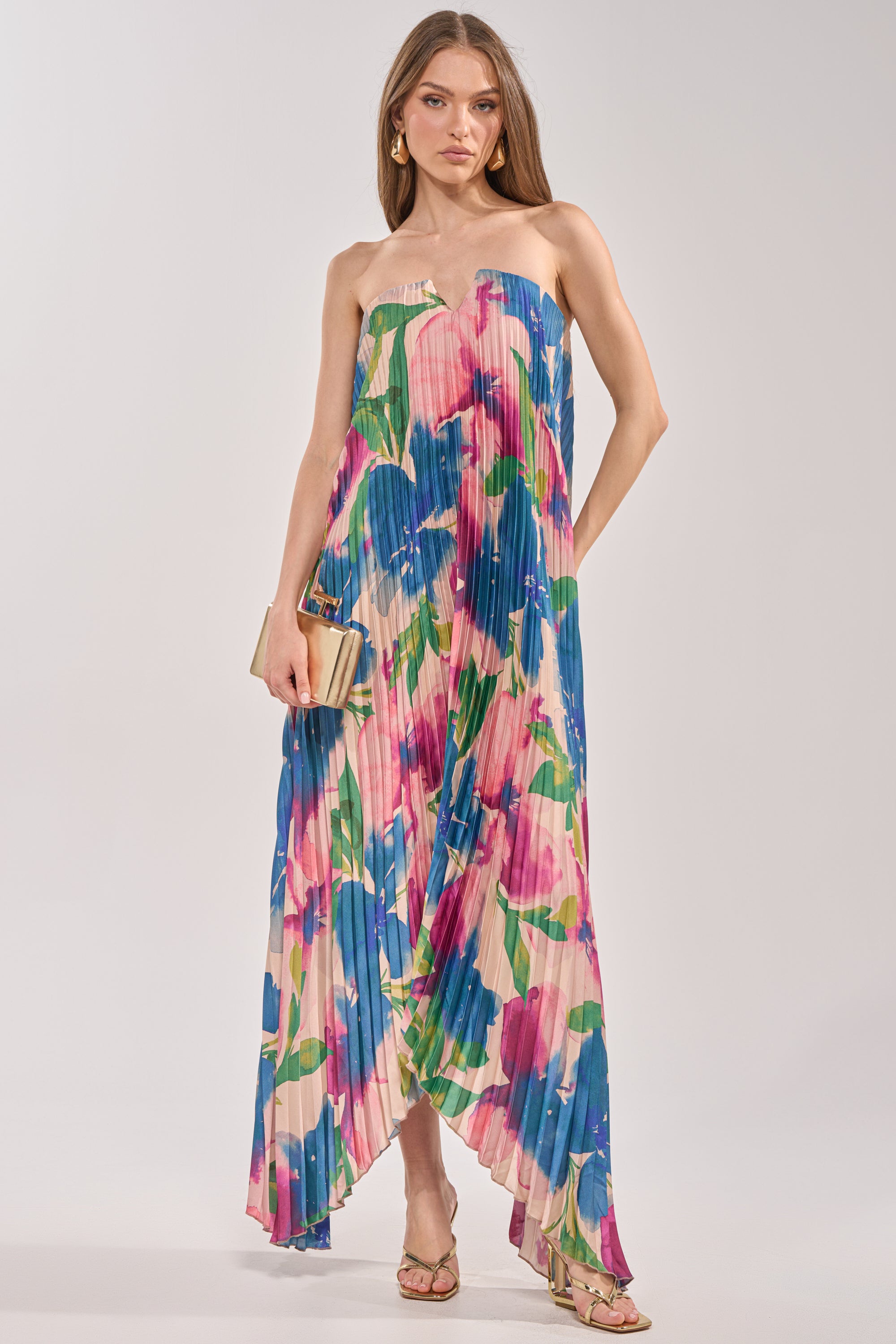 A woman wears the COCKTAIL HOUR FLORAL STRAPLESS MAXI DRESS in a blue, pink, and green floral print, paired with a beige clutch, gold heeled sandals, and hoop earrings. 0