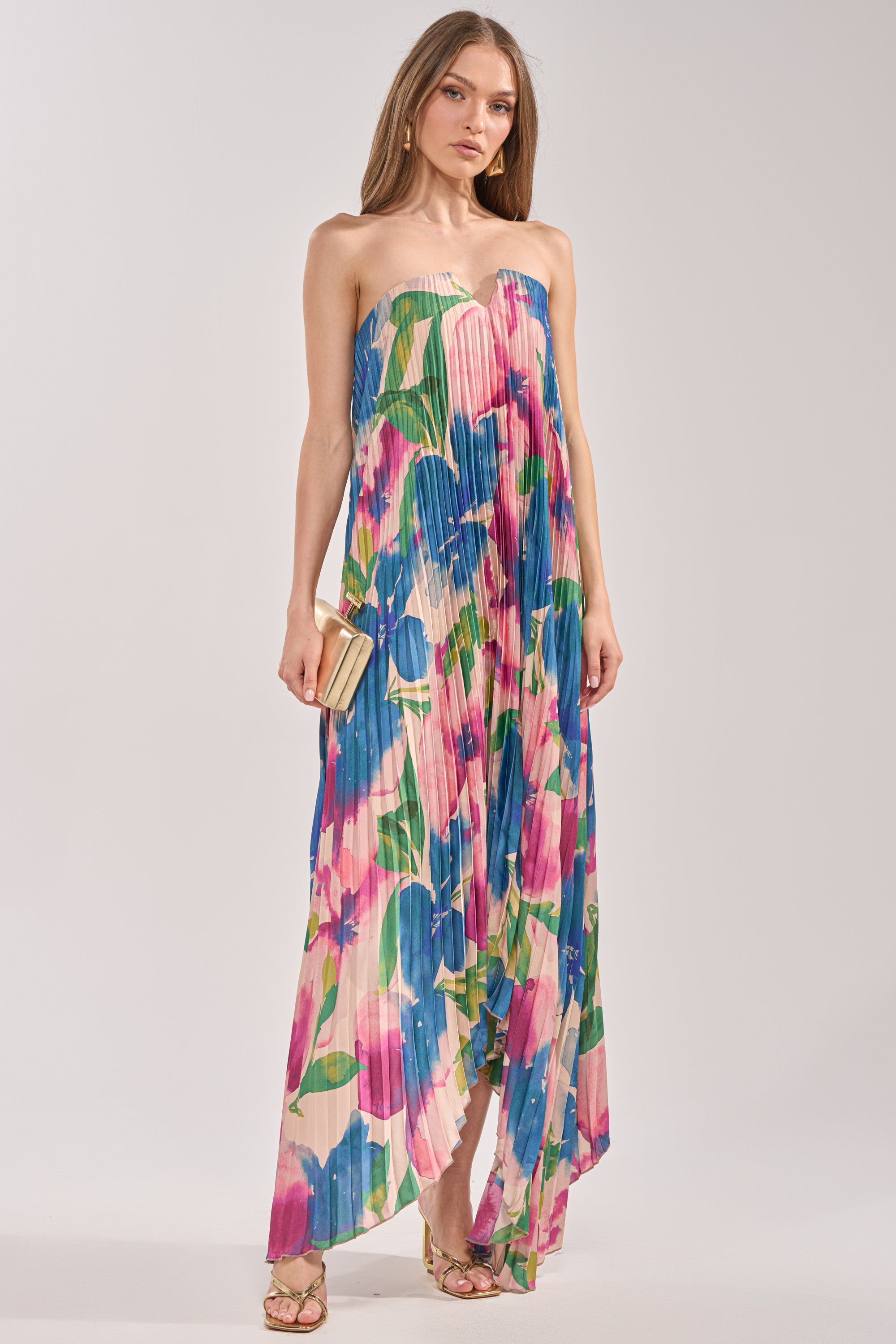 A woman in the COCKTAIL HOUR FLORAL STRAPLESS MAXI DRESS with pleated satin accents, gold sandals, and a gold clutch stands against a light background. Ideal for cocktail parties. 1