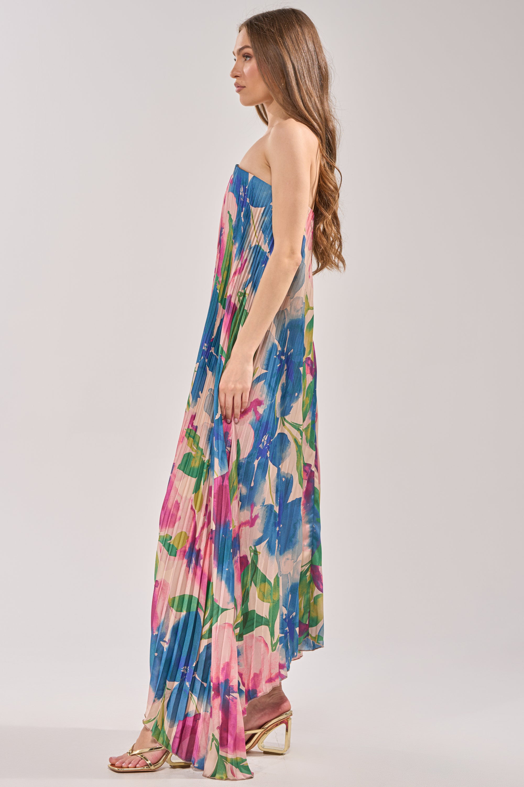 A woman with long brown hair stands in profile, wearing the COCKTAIL HOUR FLORAL STRAPLESS MAXI DRESS, featuring a floral print in blue, pink, and green hues. She pairs it with open-toe heeled sandals. 3
