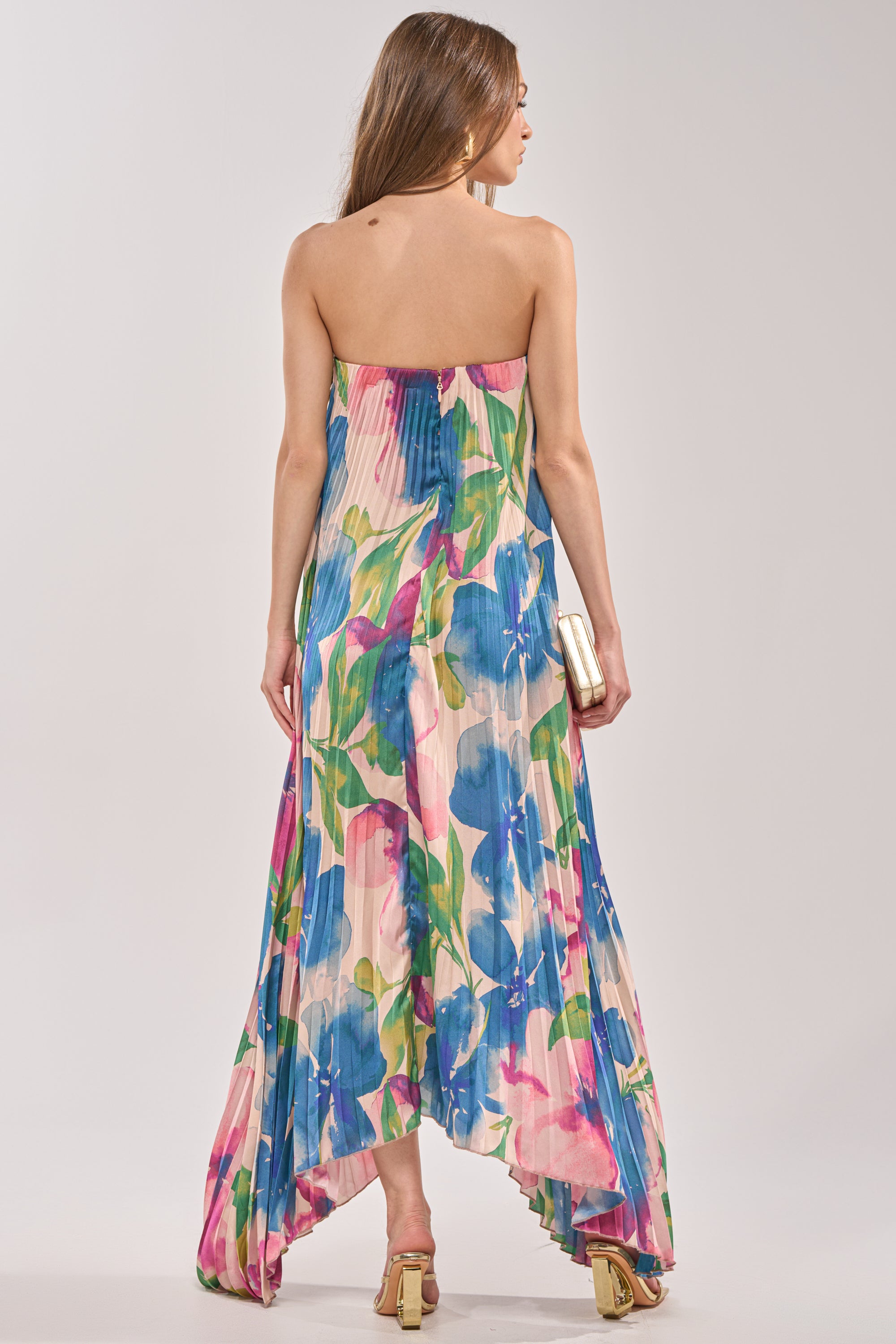 Woman stands with her back to the camera, wearing the COCKTAIL HOUR FLORAL STRAPLESS MAXI DRESS featuring a vibrant floral print and uneven hemline. She holds a small rectangular clutch and wears light-colored heeled sandals. 4