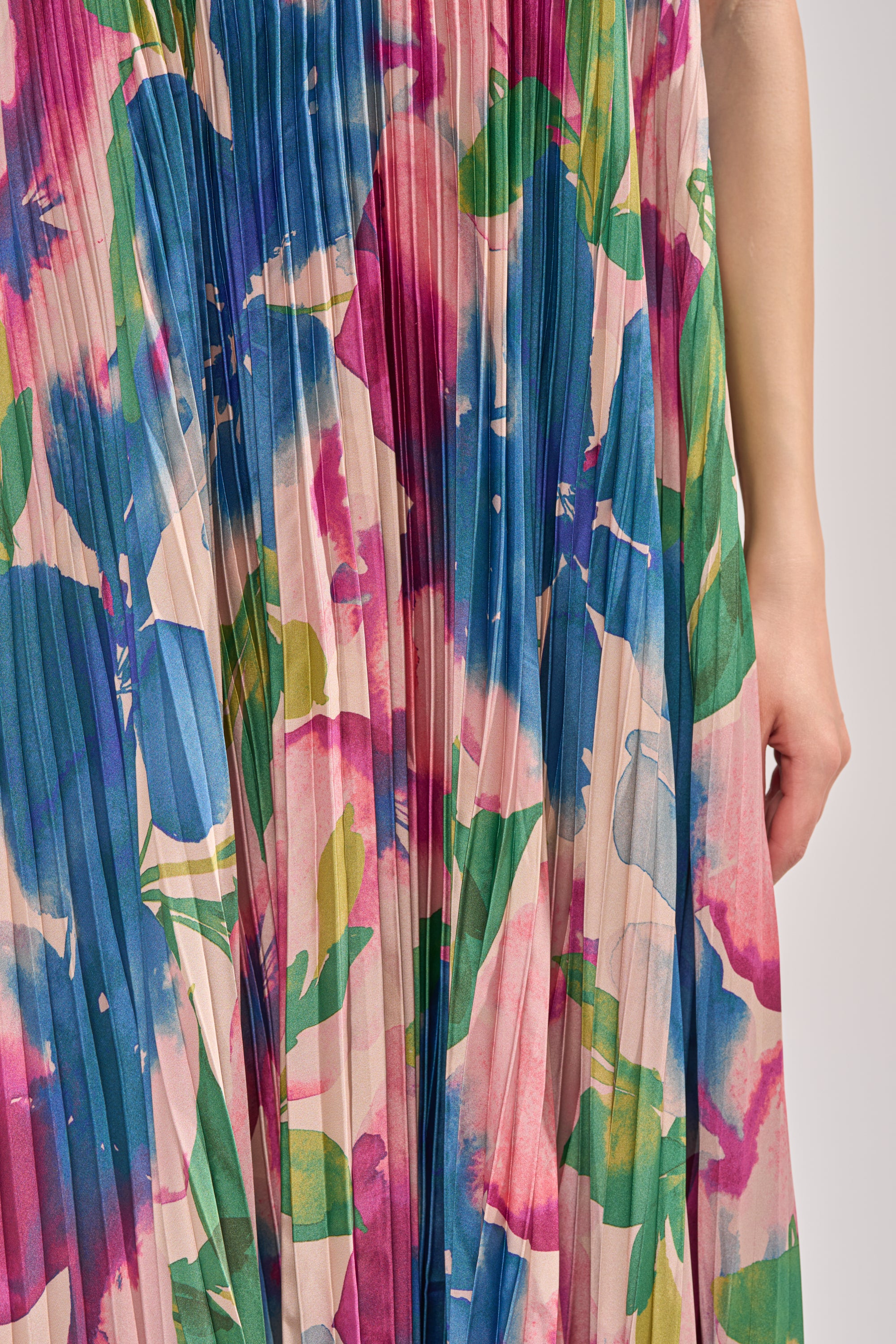 A close-up of the COCKTAIL HOUR FLORAL STRAPLESS MAXI DRESS, showing its pleated skirt with an abstract floral pattern in pink, green, blue, and beige. A person's arm and hand appear on the right side. 5