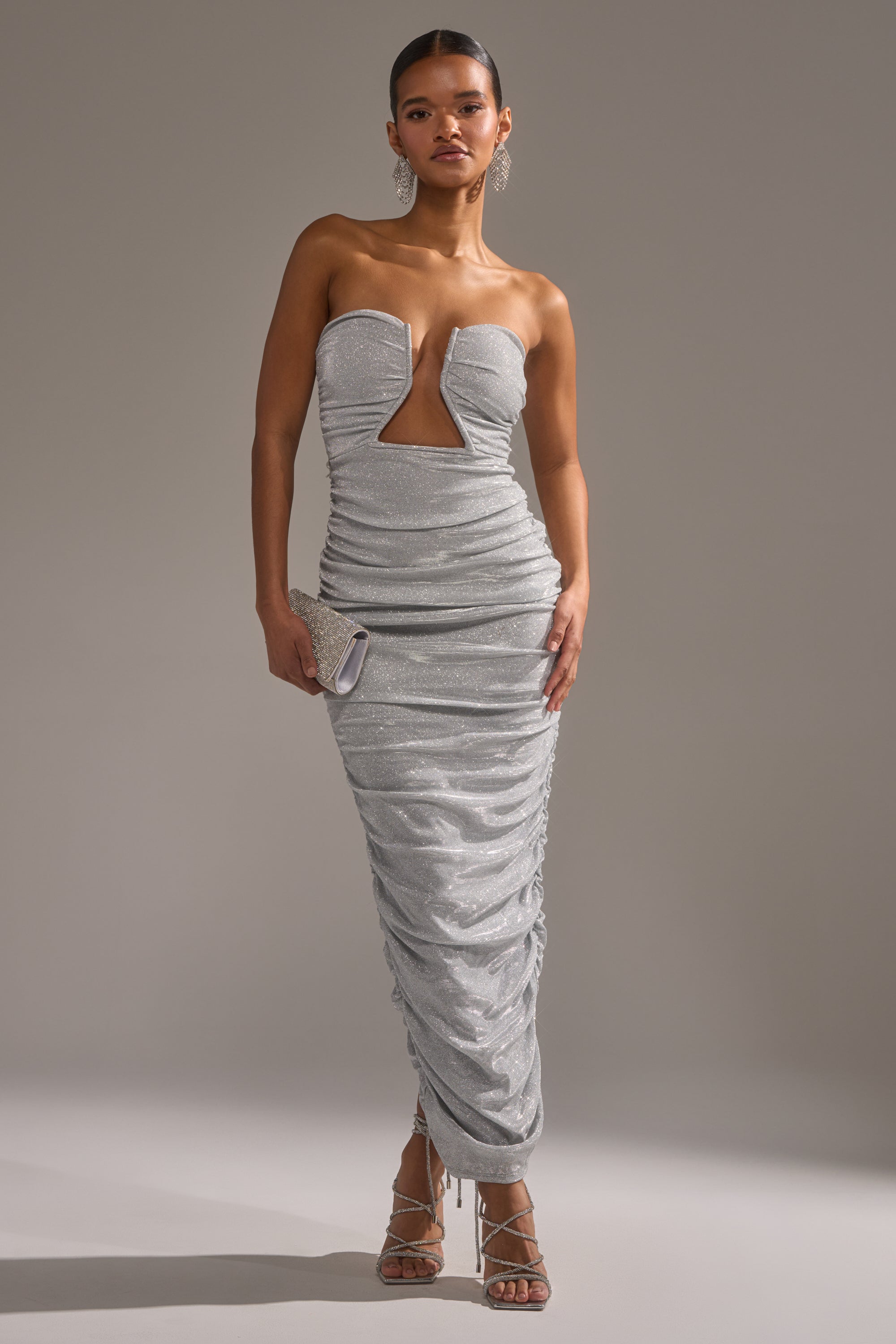 A woman models the CANT GET ENOUGH SLINKY RUCHED MAXI DRESS, featuring a strapless, glitter design with ruching and a front cutout. She completes the look with strappy heels, statement earrings, and a silver clutch against a gray backdrop.