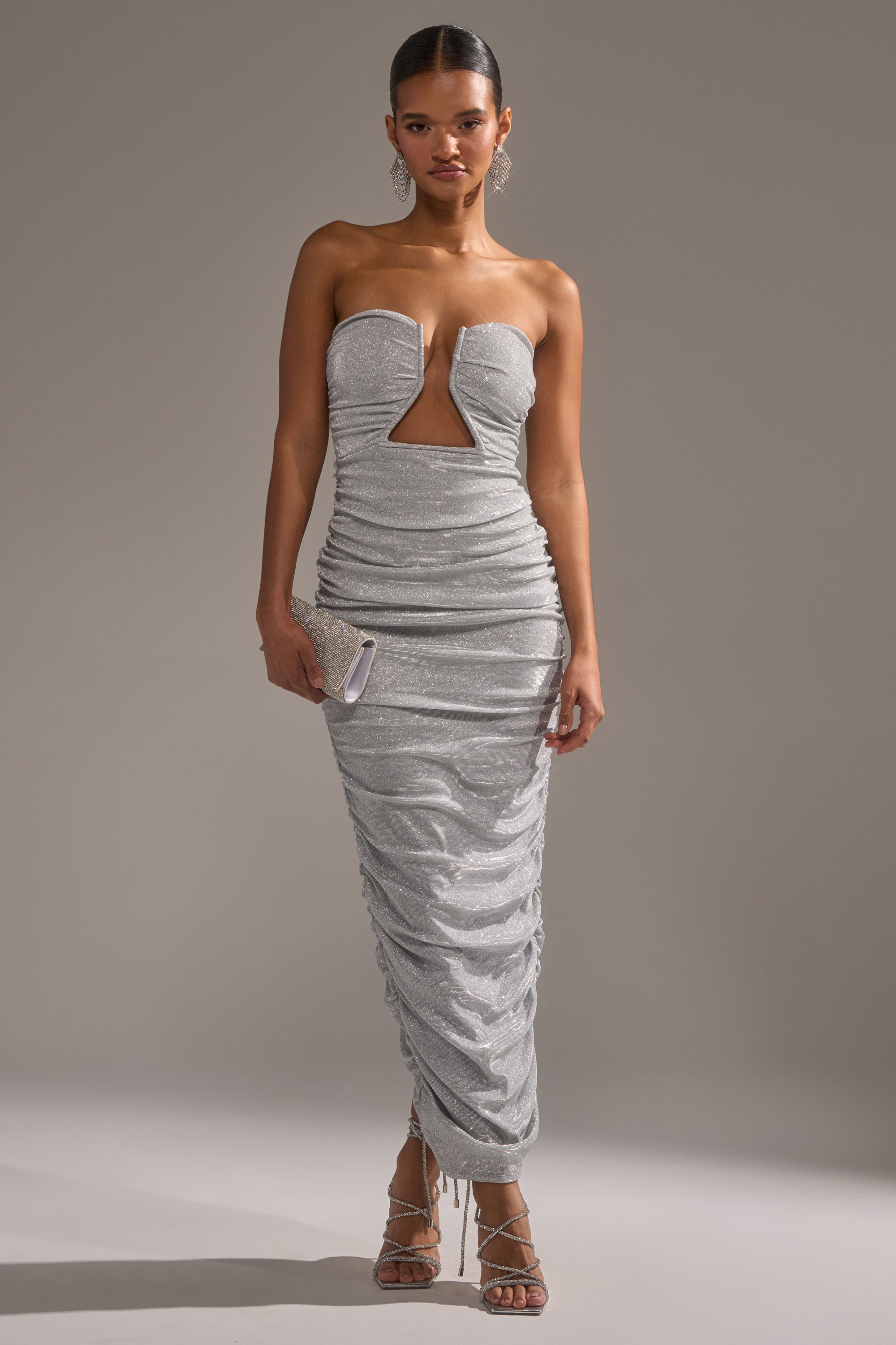 A woman wears the CANT GET ENOUGH SLINKY RUCHED MAXI DRESS with a silver clutch and strappy sandals, styled against a plain gray background.