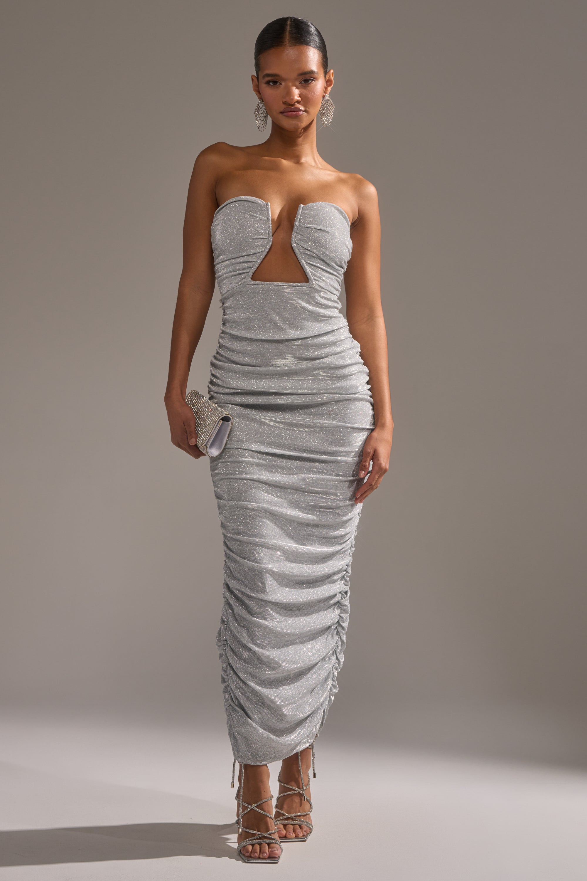 A woman models the CANT GET ENOUGH SLINKY RUCHED MAXI DRESS, a strapless silver glitter gown with shimmery ruched details and a front cutout. She pairs it with strappy heels, a small clutch, slicked-back hair, against a neutral background.