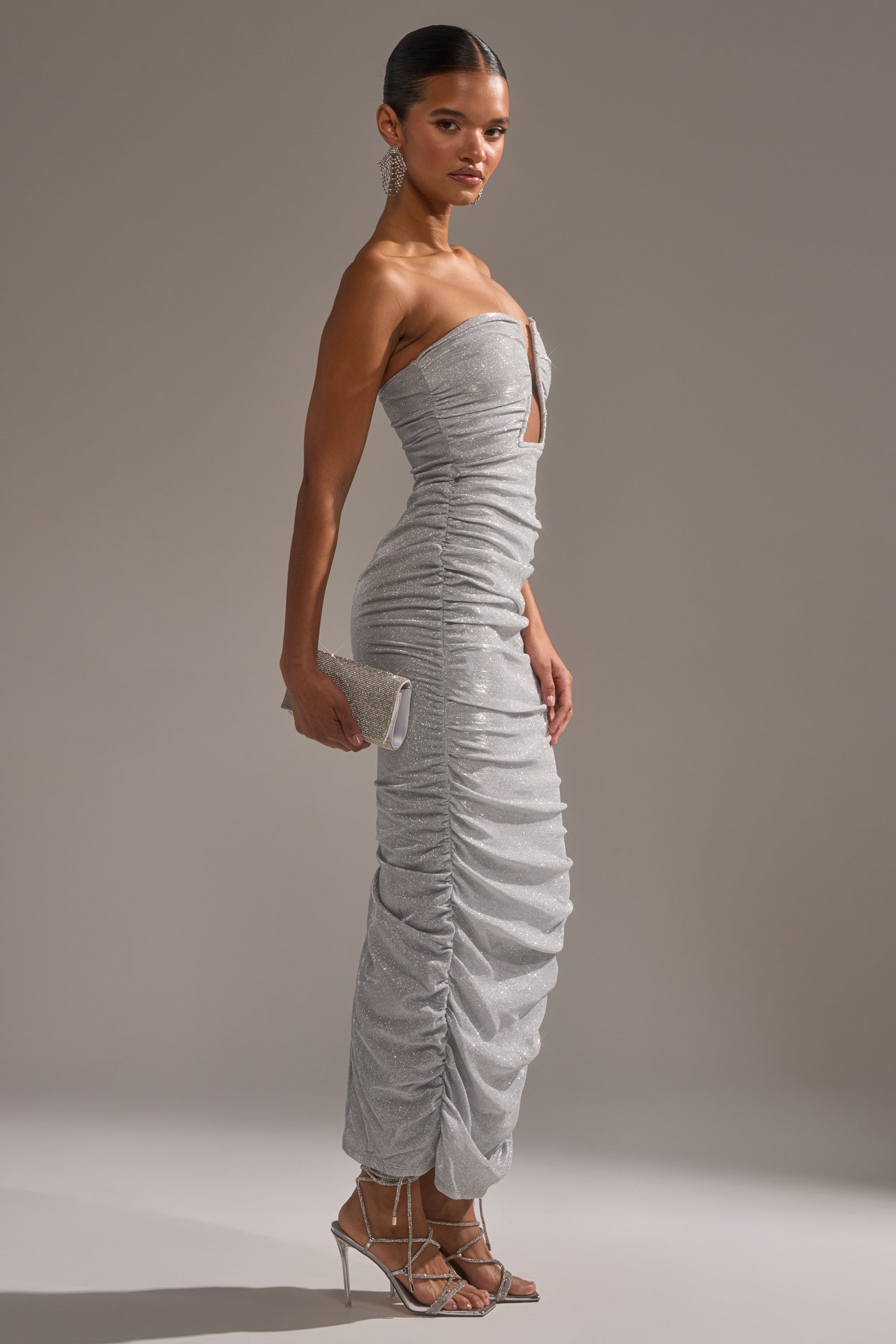 A woman models the CANT GET ENOUGH SLINKY RUCHED MAXI DRESS, standing sideways with a matching clutch. She pairs the strapless dress with strappy silver heels, bold dangling earrings, and sleeked-back hair against a plain gray background.