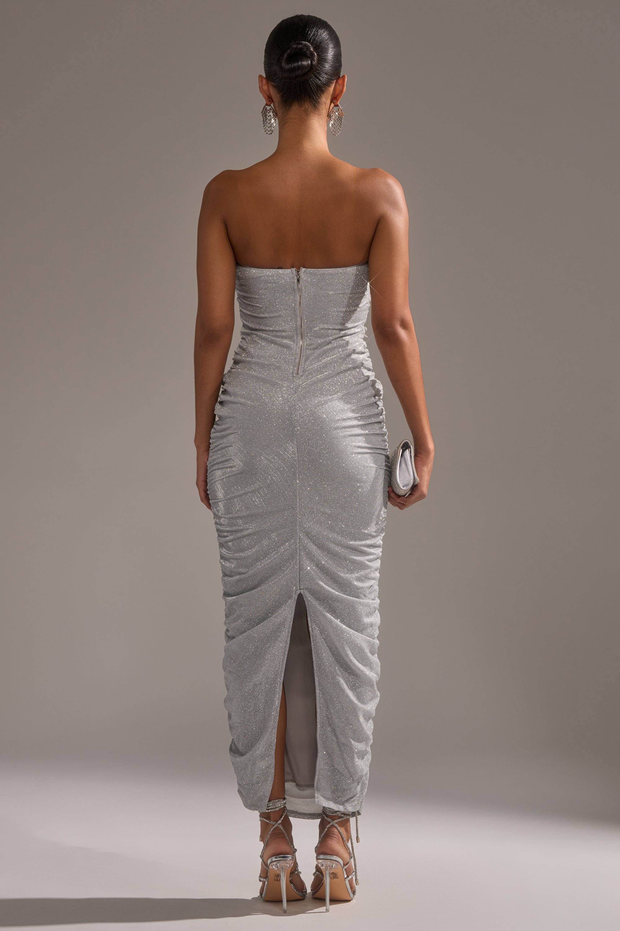 A woman faces away, wearing the CANT GET ENOUGH SLINKY RUCHED MAXI DRESS with a back slit. She holds a small clutch, wears silver heels and statement earrings, and her hair is styled in a sleek bun.