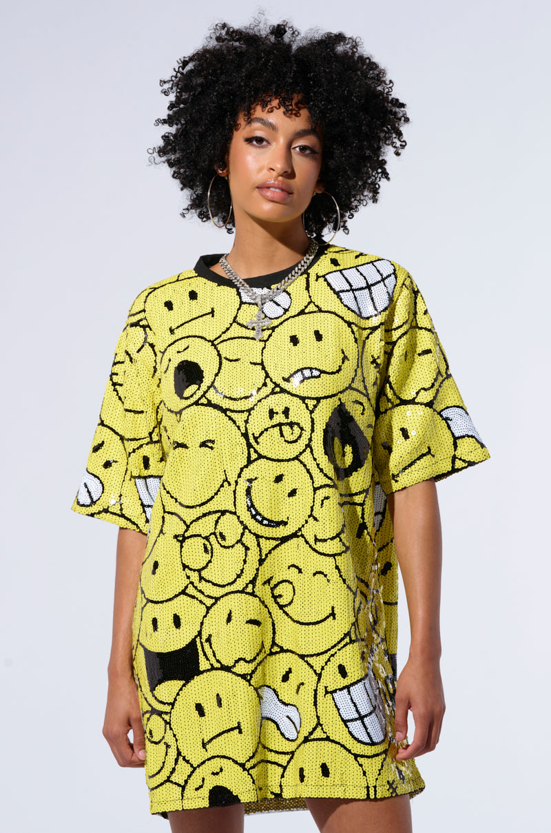 A person with curly hair is wearing the loose-fitting, short-sleeved SMILE FOR SPARKLES T SHIRT in bright yellow, each smiley face displaying different emotions. The background is plain and light-colored.