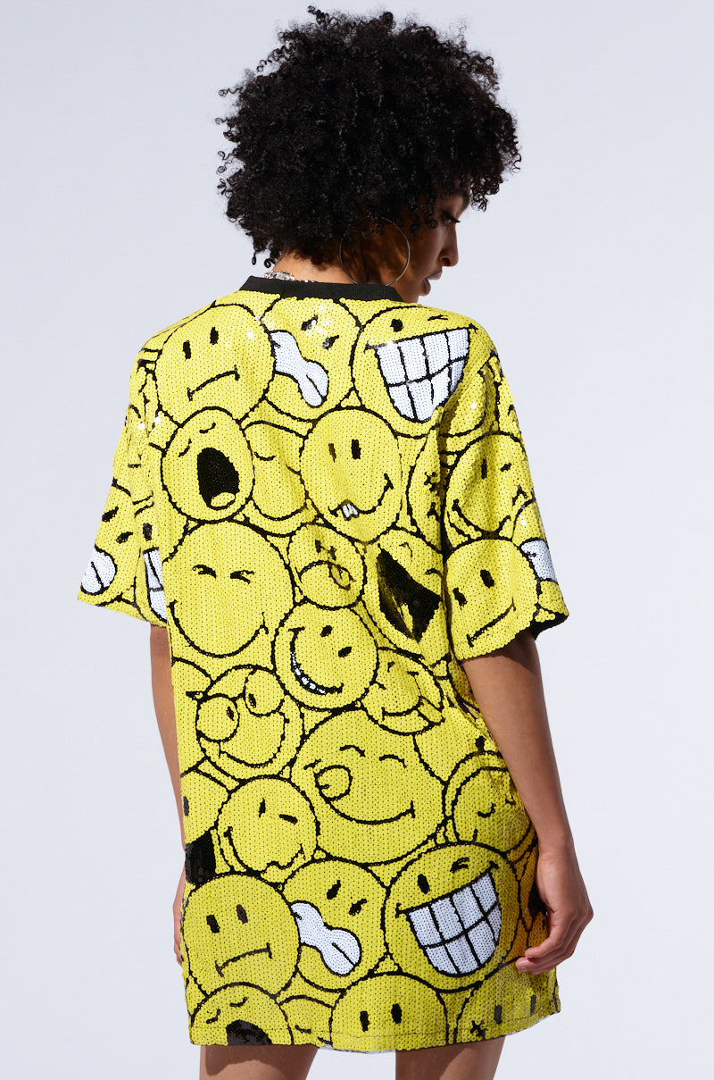 A person with curly hair stands facing away, wearing an oversized "SMILE FOR SPARKLES" T-shirt adorned with various expressive smiley faces. The background is plain and light-colored.