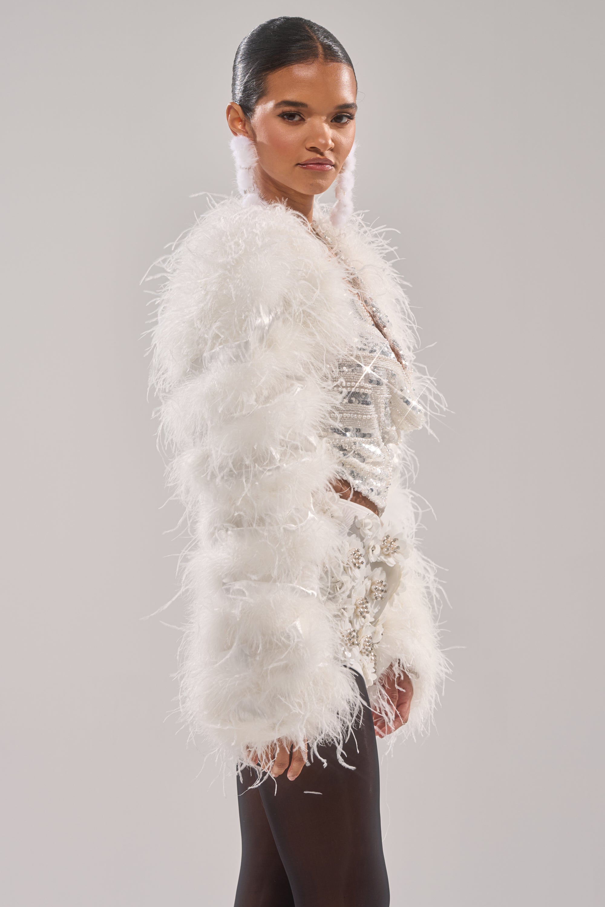 A woman models the PARK AVE FEATHER BOLERO in white with long sleeves, layered over a shimmering silver outfit and black tights, her hair slicked back, set against a simple light gray background.
