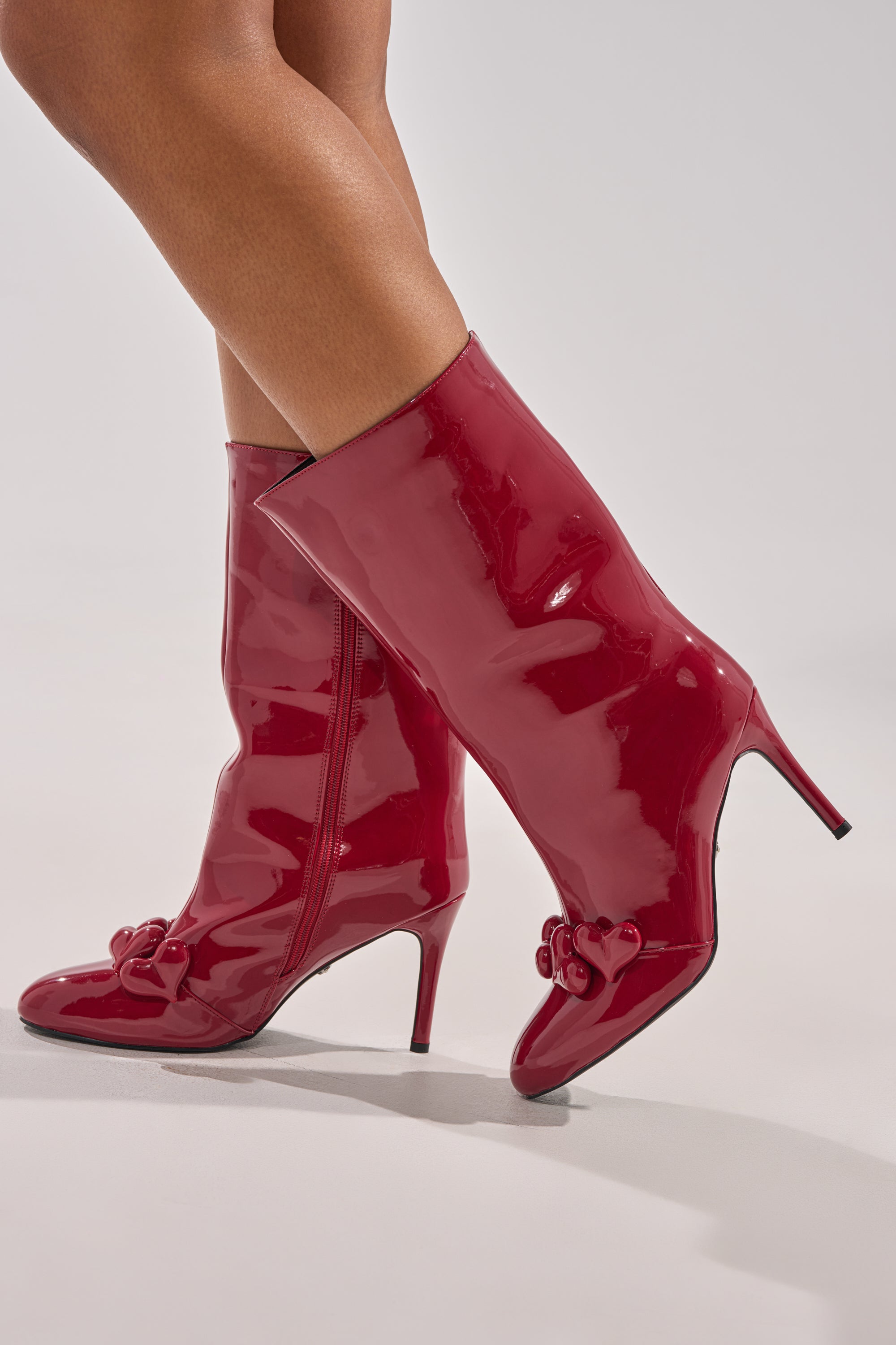 Model wearing AZALEA WANG FEDELE Red Patent Heart Embellished Bootie with stiletto heels, standing against a plain light background.